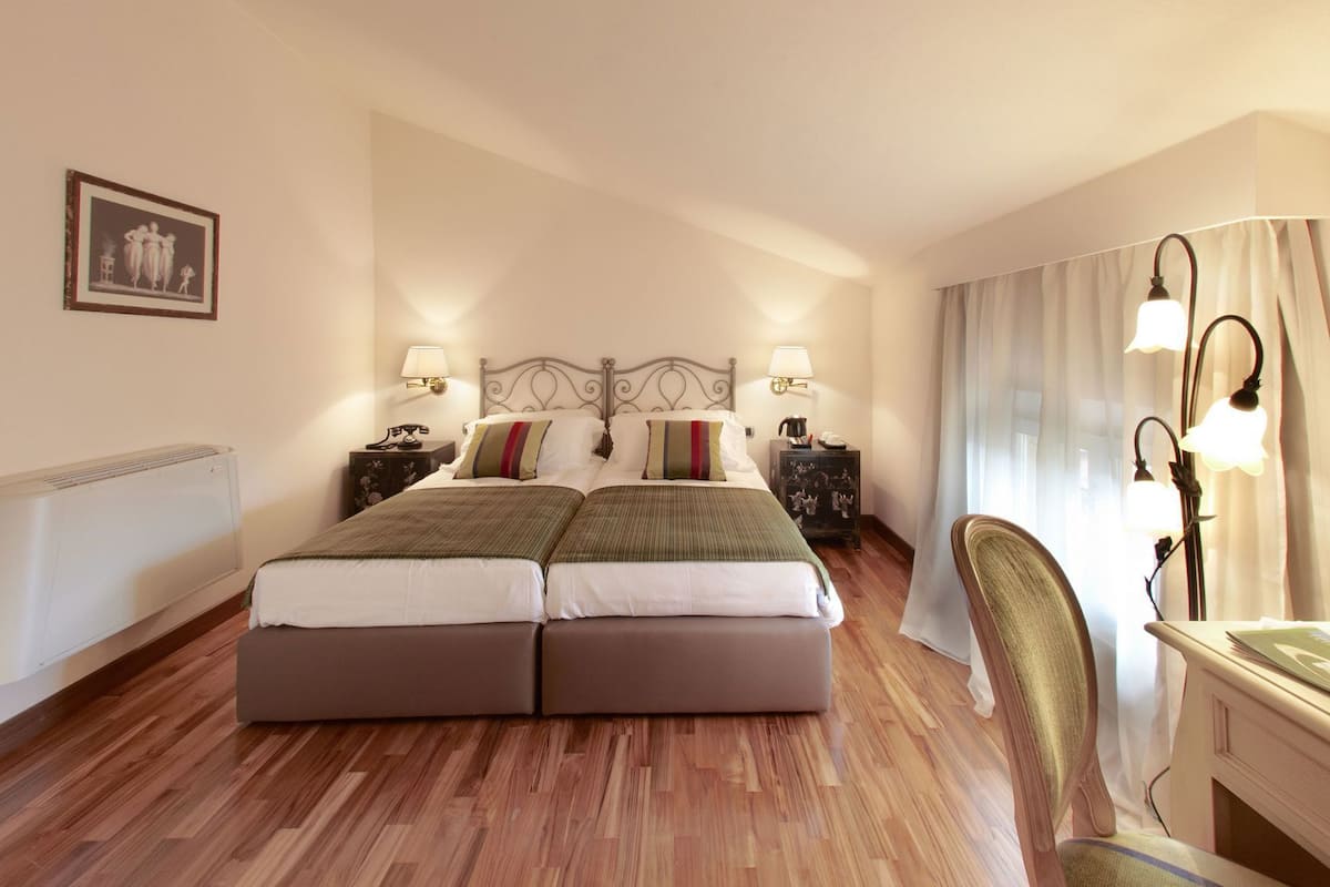 A double bed with a headboard, two bedside tables with lamps, a chair, a radiator, and a framed picture on the wall.