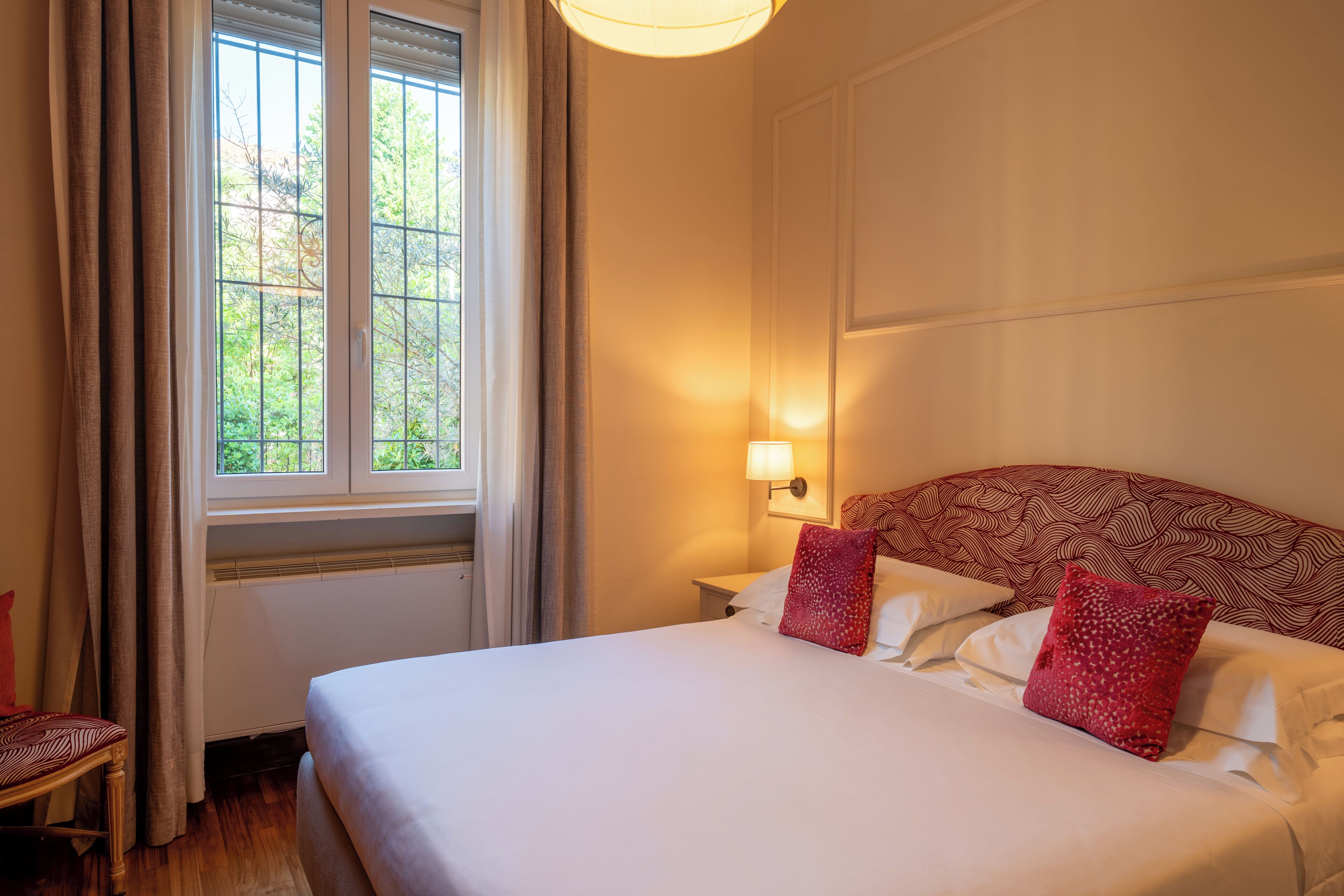 A hotel room with a bed, a window with curtains, a nightstand with a lamp, and a patterned headboard.