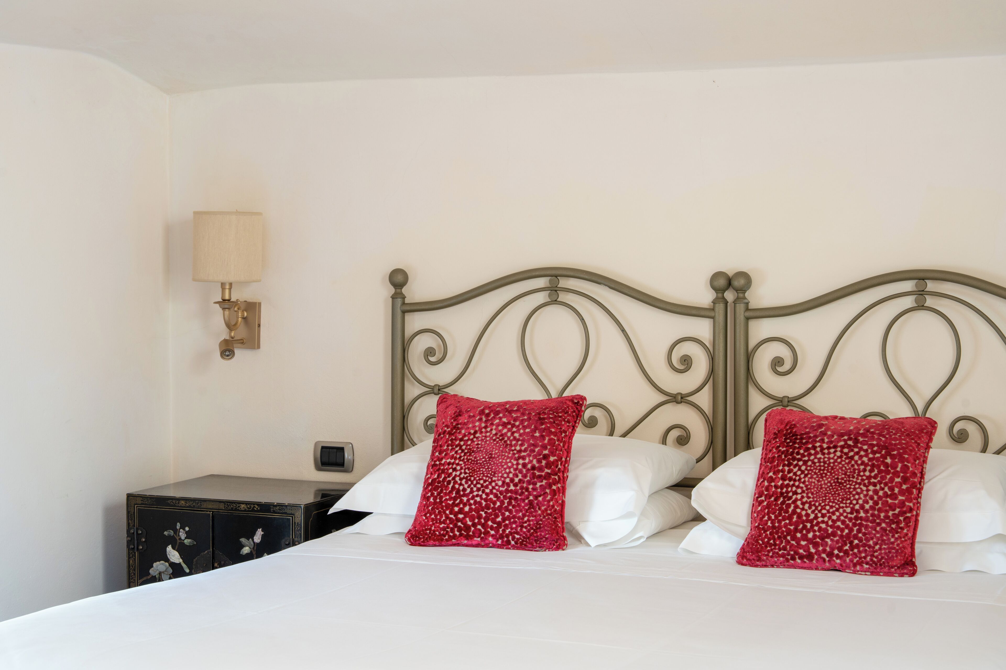 Ein Bett mit Metallkopfteil, zwei rote Kissen, ein Nachttisch und eine Wandlampe.
