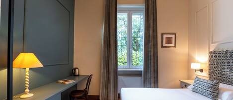 Une chambre d’hôtel comprenant un lit, un bureau, une chaise, une fenêtre avec des rideaux et une lampe fixée au mur.
