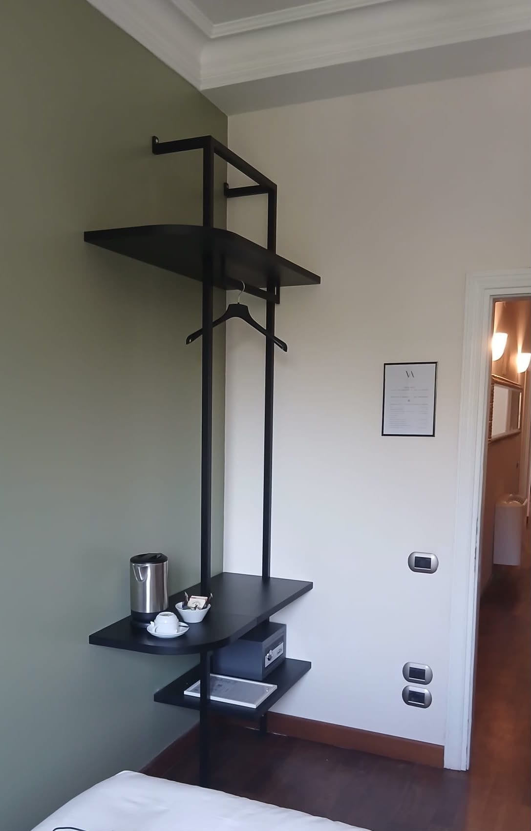 Una habitación con una estantería metálica negra contra la pared, una puerta blanca con un aviso enmarcado y un cubo de basura en la esquina.