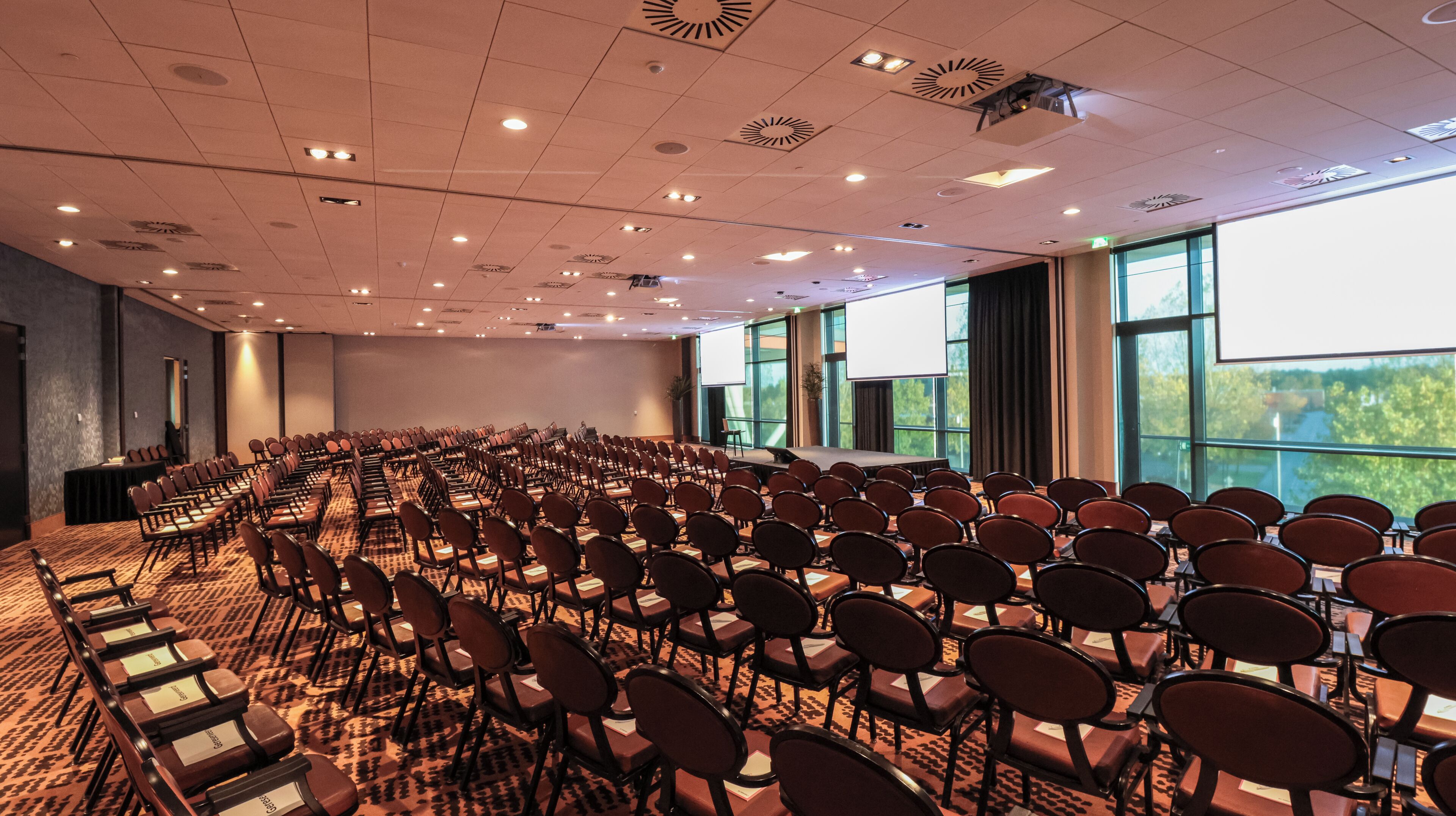 Un salón de conferencias con filas de sillas, un escenario y ventanas amplias.