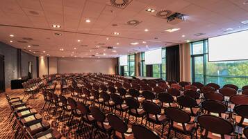 Un salón de conferencias con filas de sillas, un escenario y ventanas amplias.