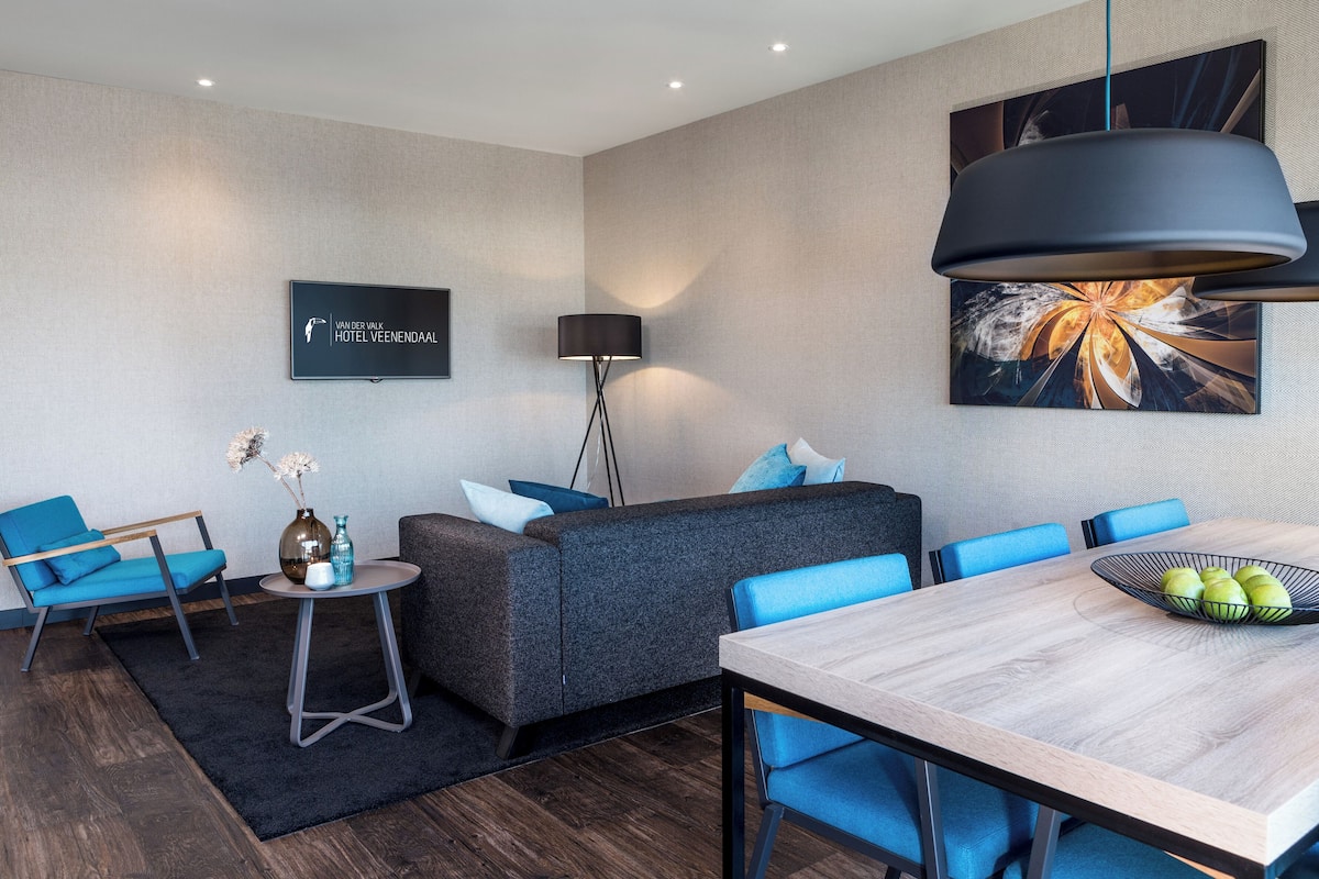 A modern hotel room with a grey sofa, blue chairs, a wooden dining table, and a black floor lamp.