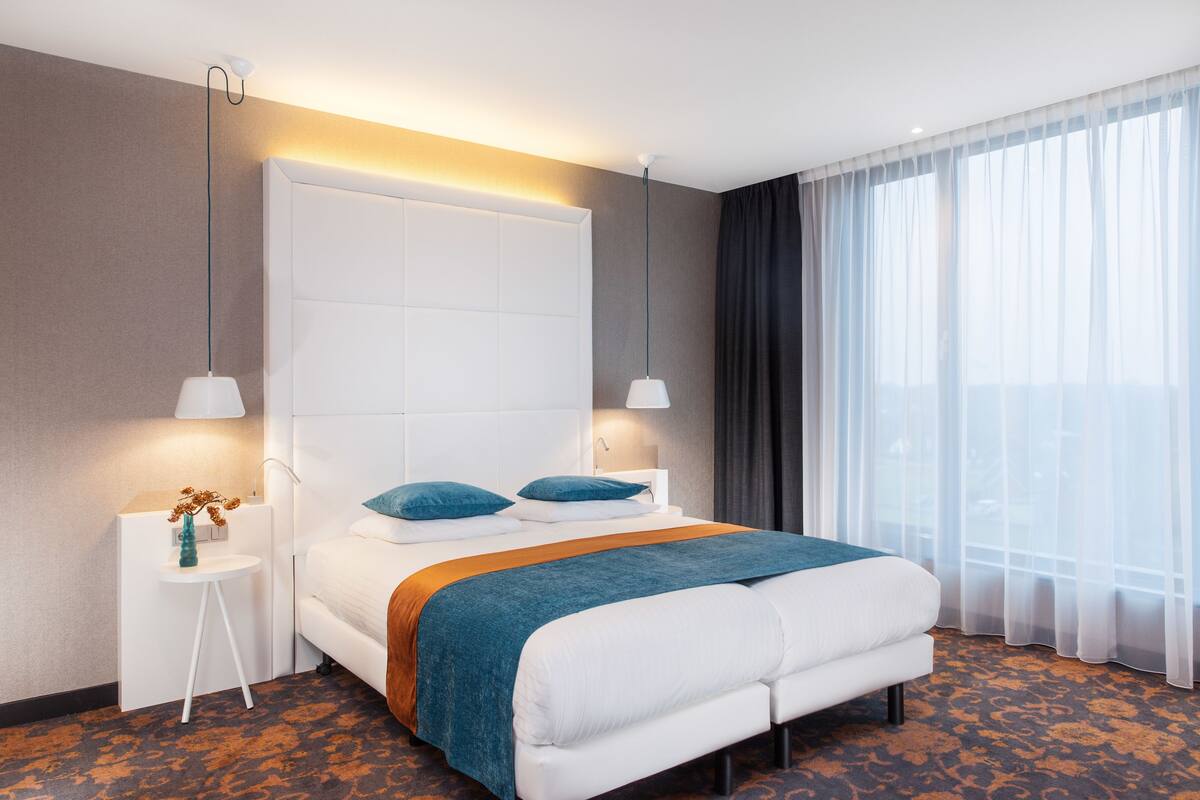 A modern hotel room with a large bed, two bedside lamps, a small side table with a vase, and a large window with sheer curtains.