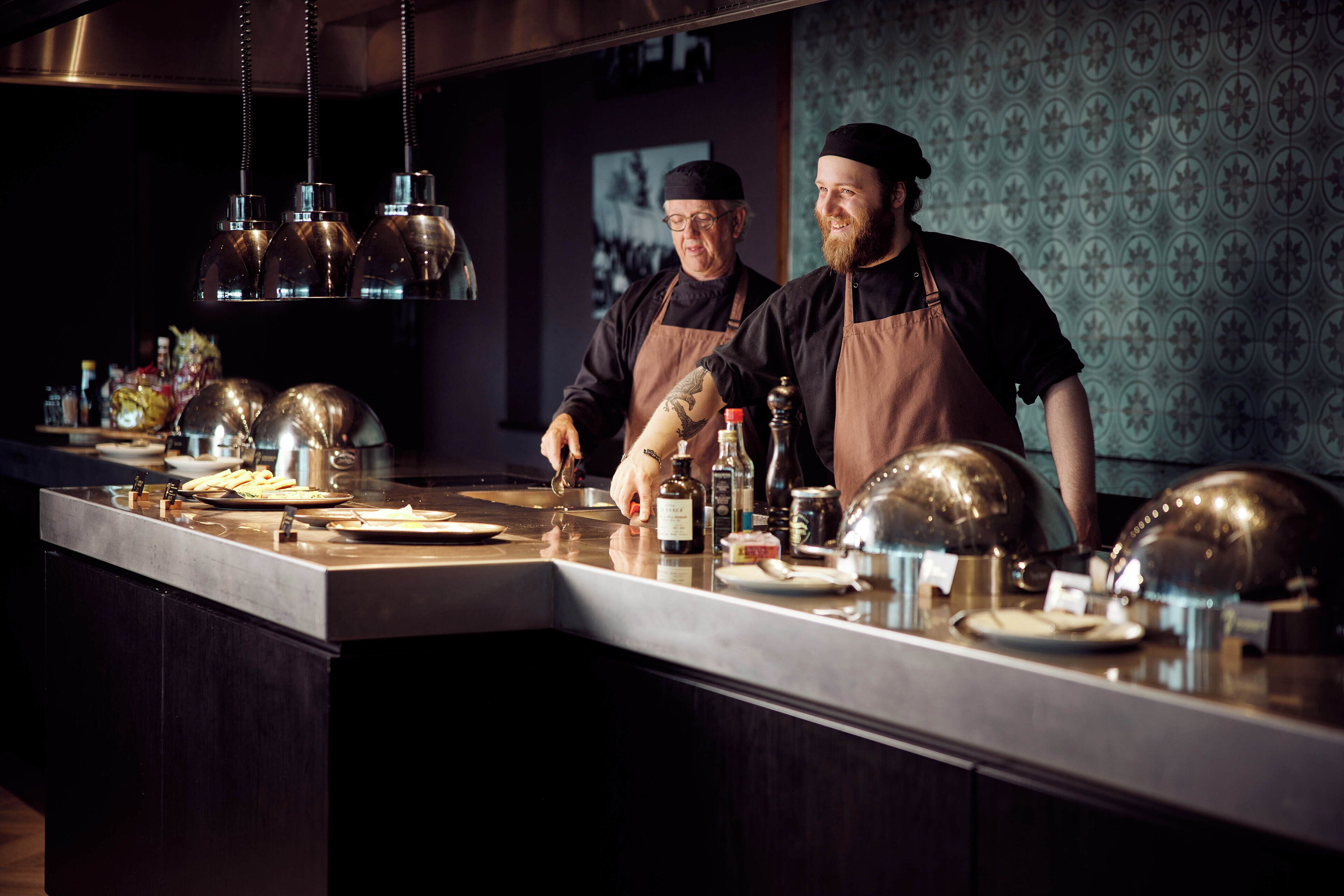 A kitchen counter with chefs preparing food, various cooking utensils, and ingredients.