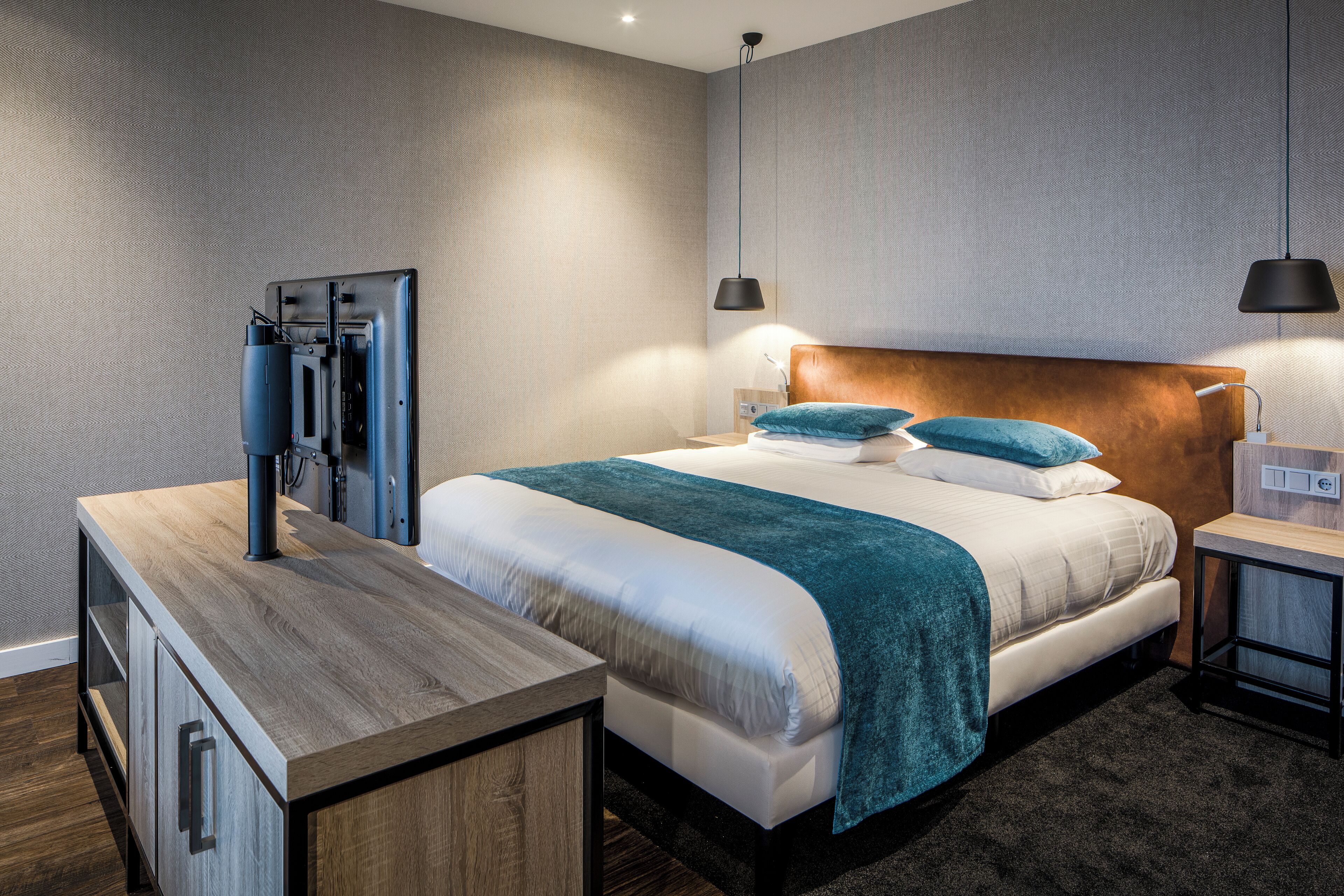 Habitación de hotel moderna con una cama grande, una mesita de noche de madera, un televisor y dos lámparas colgantes.
