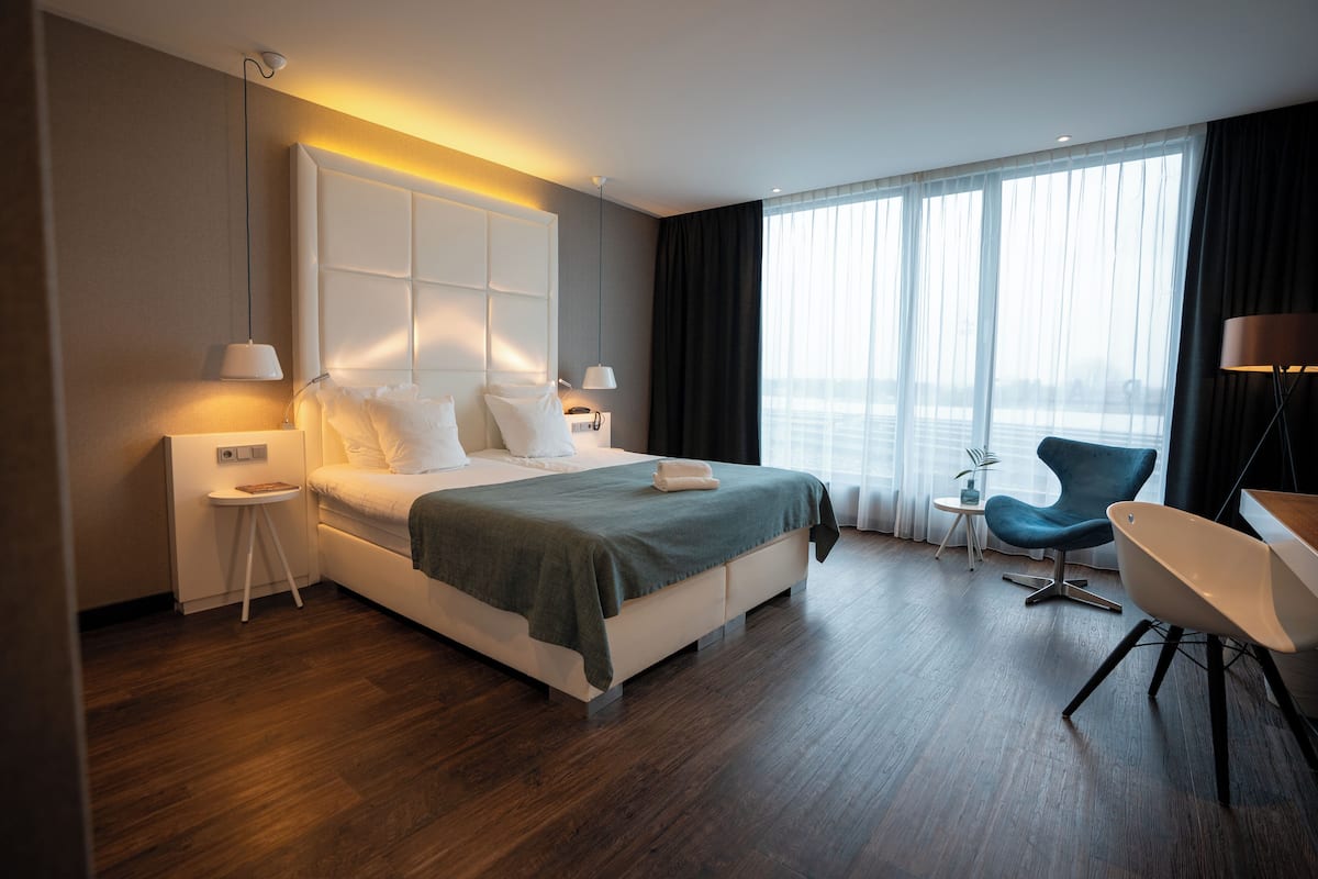 A modern hotel room with a large bed, bedside tables, a desk, a chair, and a view of the city through large windows.