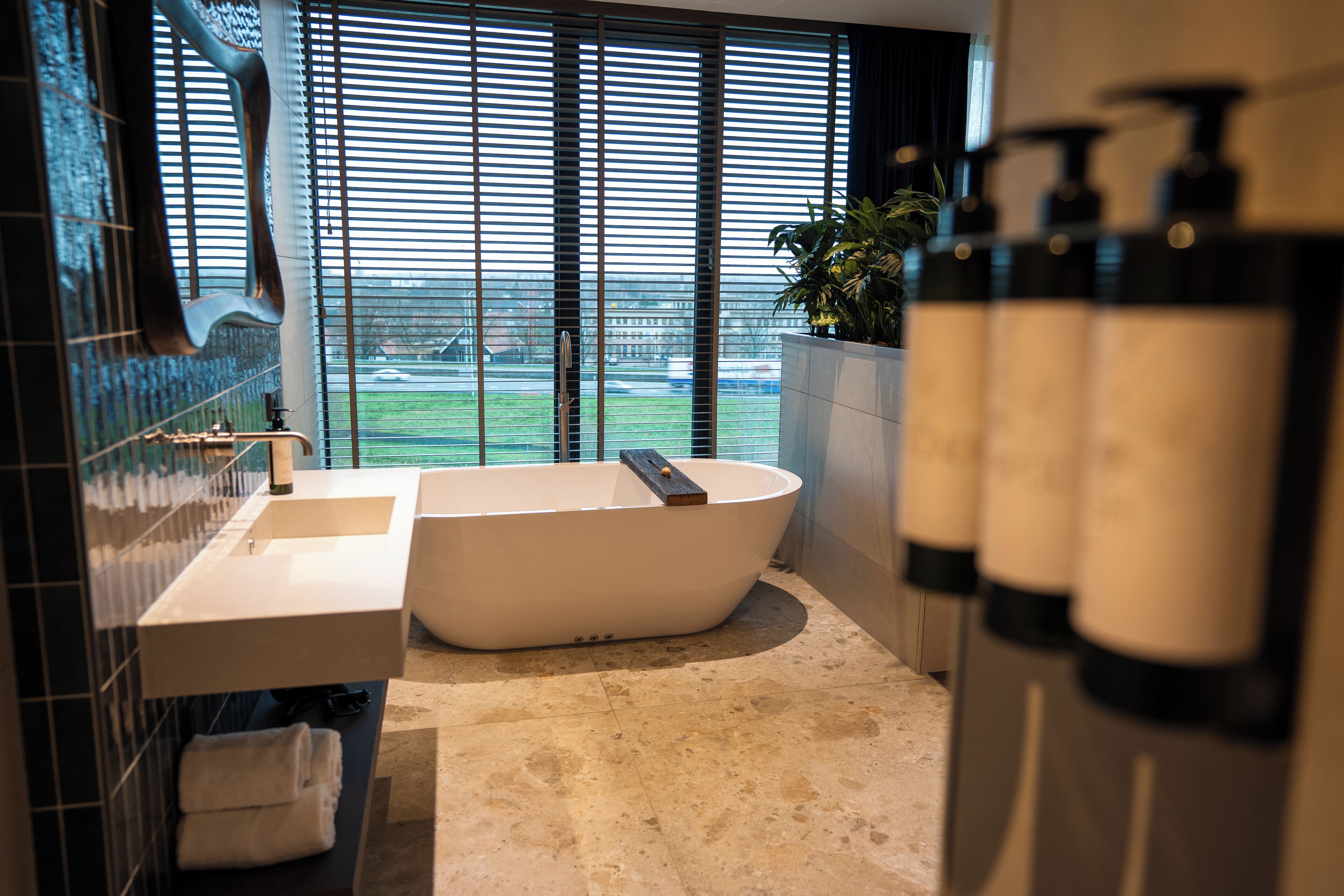 Een moderne badkamer met een vrijstaand bad, een wastafel en een groot raam met jaloezieën.