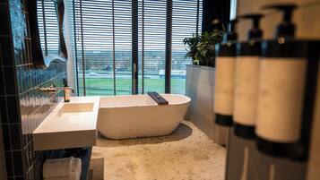 Een moderne badkamer met een vrijstaand bad, een wastafel en een groot raam met jaloezieën.