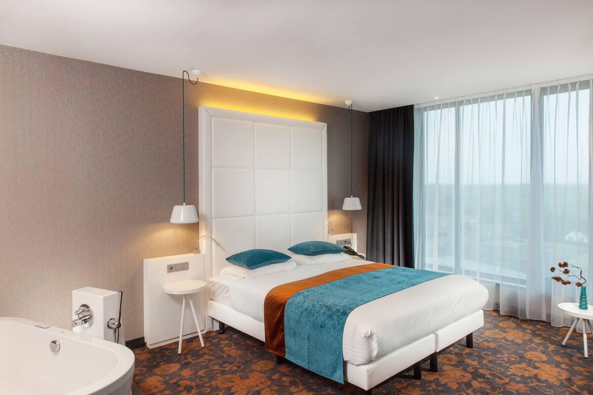 A modern hotel room with a large bed, a bedside table, a nightstand, a white bathtub, and a window with curtains.