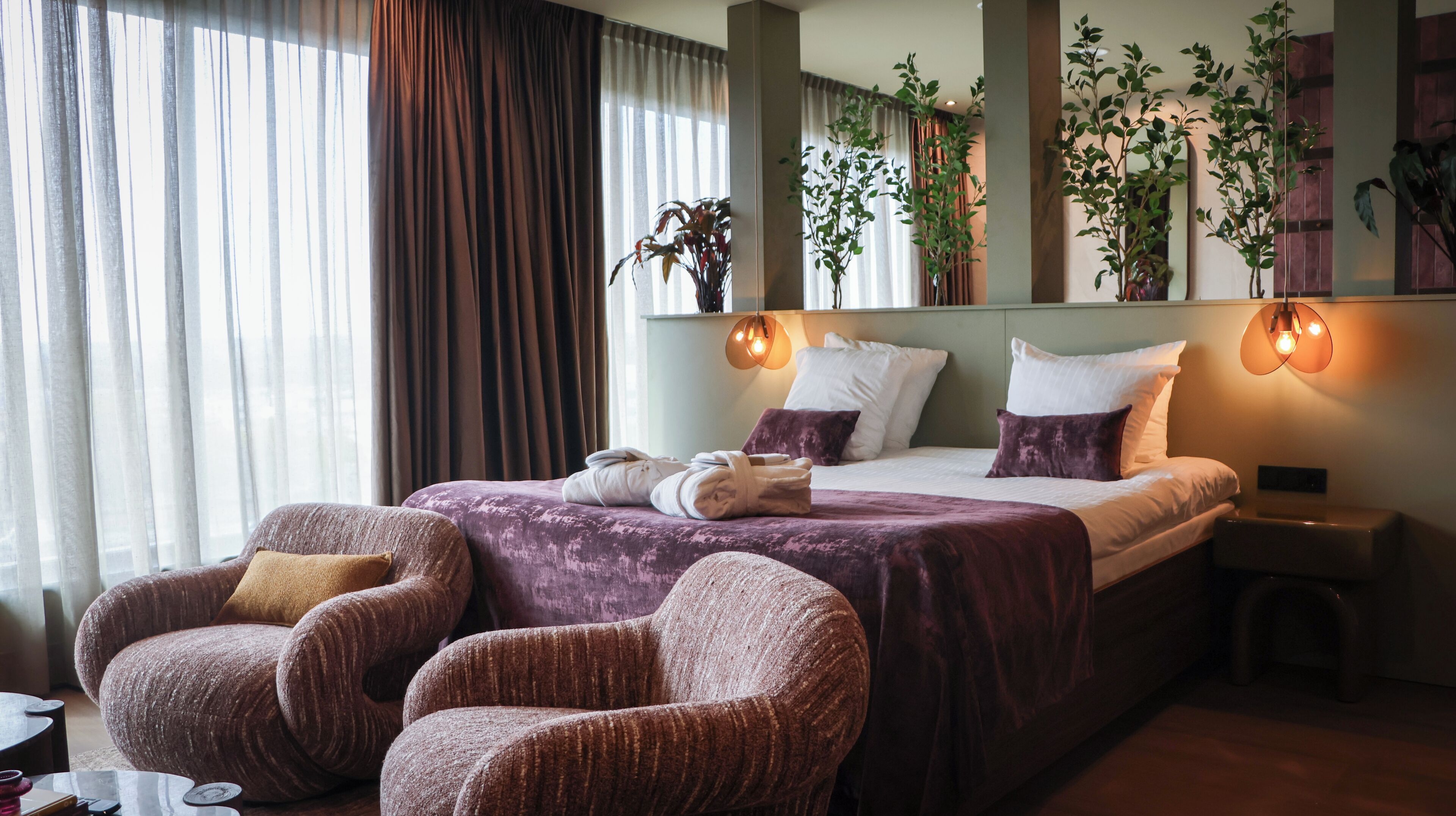 Hotelkamer met een bed, twee fauteuils, een nachtkastje en een spiegel met planten.