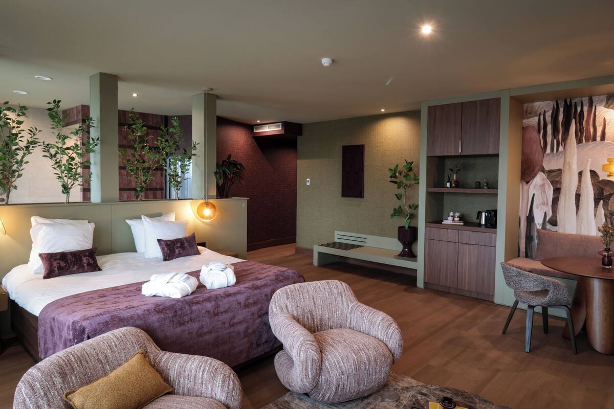 A modern hotel room with a bed, two armchairs, a small table, a bench, a dining area, and a wall with a mural.