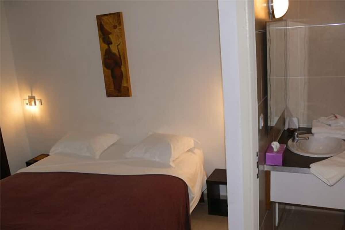 A hotel room with a bed, a nightstand, a wall-mounted artwork, and a bathroom with a sink and mirror.