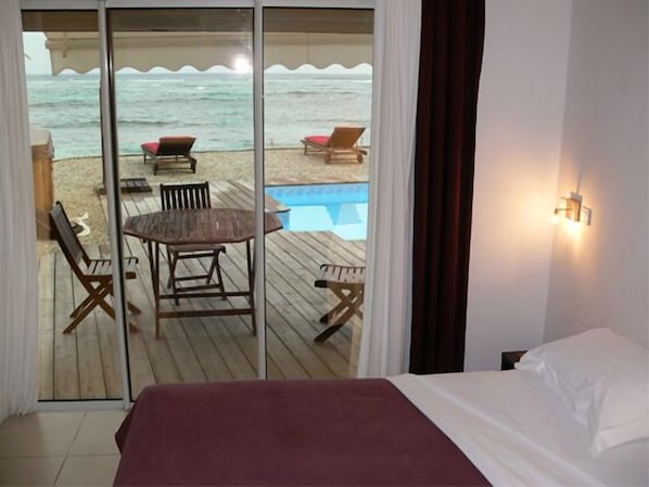 Una habitación con una cama, una mesa de madera con sillas, vista a la playa y al océano a través de un ventanal, y una piscina.