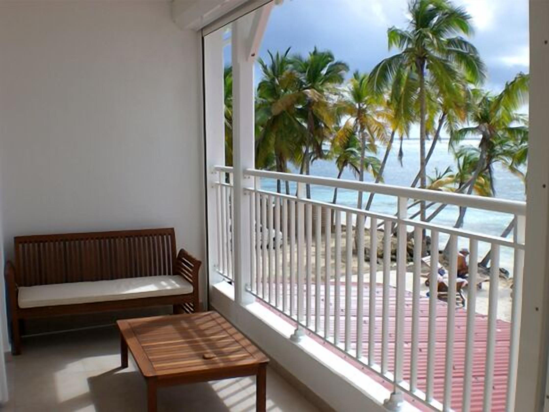 Een balkon met een houten bankje en tafel, met uitzicht op een strand met palmbomen en een heldere lucht.