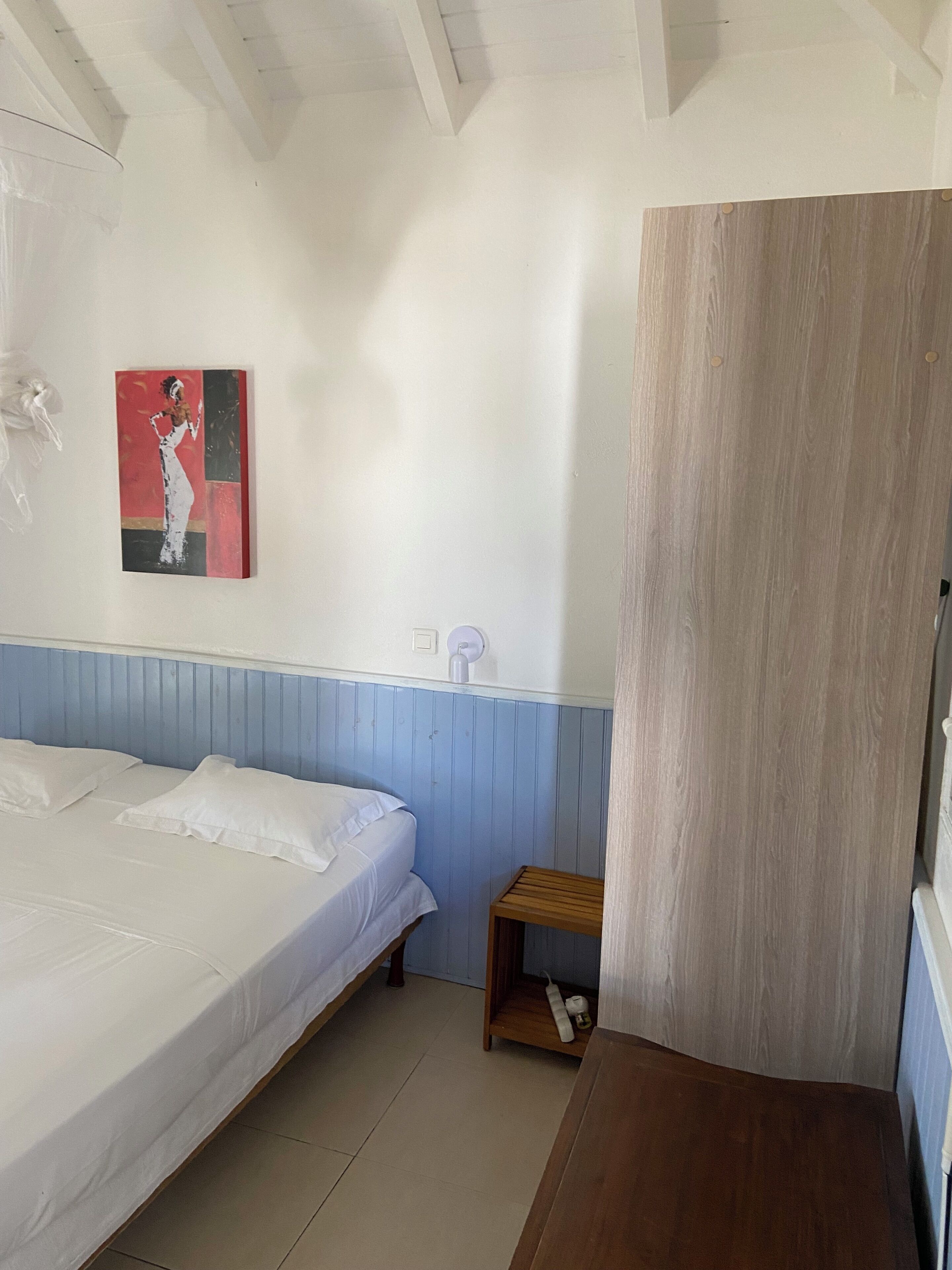 A bedroom with a bed, a small wooden nightstand, a wall-mounted light, and a painting.