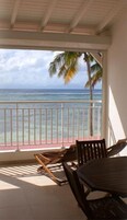 Un área de estar al aire libre techada con vista al océano, una palmera, y una mesa y sillas de madera.