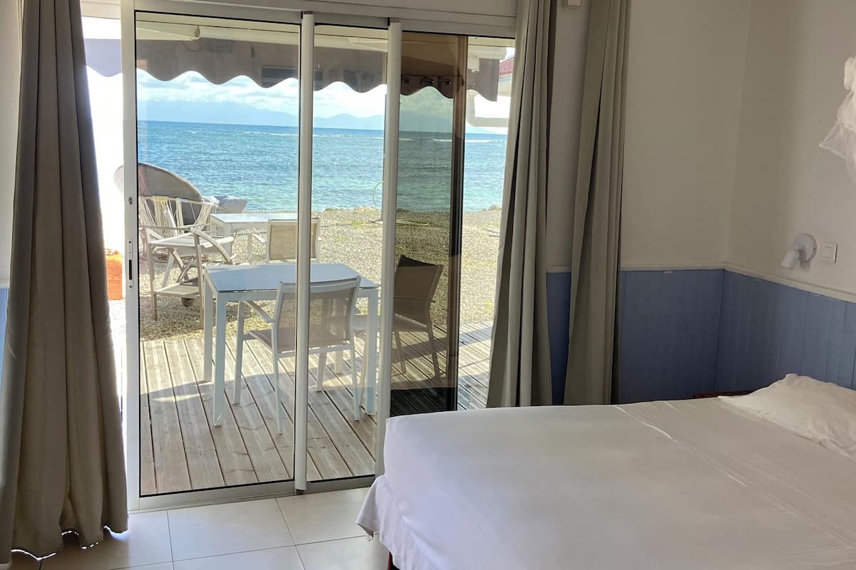 A hotel room with a bed, a sliding glass door leading to a deck with chairs and a table, and a view of the sea.