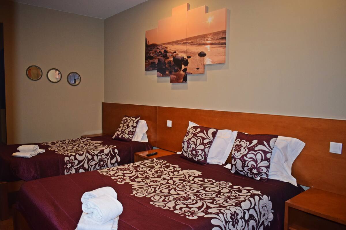 Een hotelkamer met twee bedden, elk voorzien van een beddekleed met patroon en kussens, een wanddecoratie met een strandtafereel, en drie ronde spiegels aan de muur.