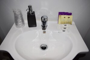 A white bathroom sink with a chrome faucet, a black soap dispenser, and a set of toiletries including a purple and yellow box labeled "NUSCH GEL SHOWER GEL". - Residencial Belo Horizonte (Porto)