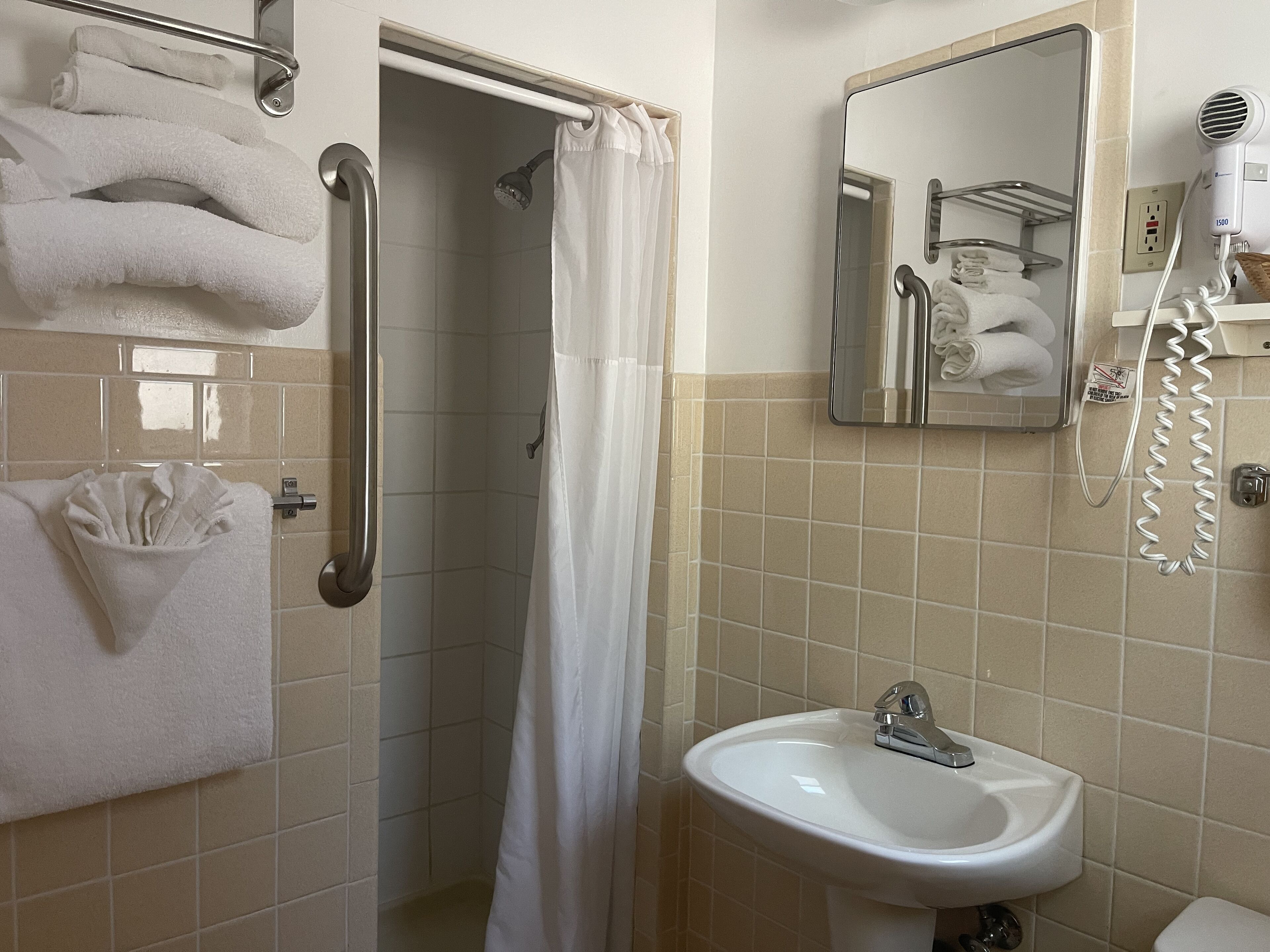 Ein Badezimmer mit Dusche, Waschbecken und Spiegel. Handtücher und ein Föhn befinden sich an der Wand.