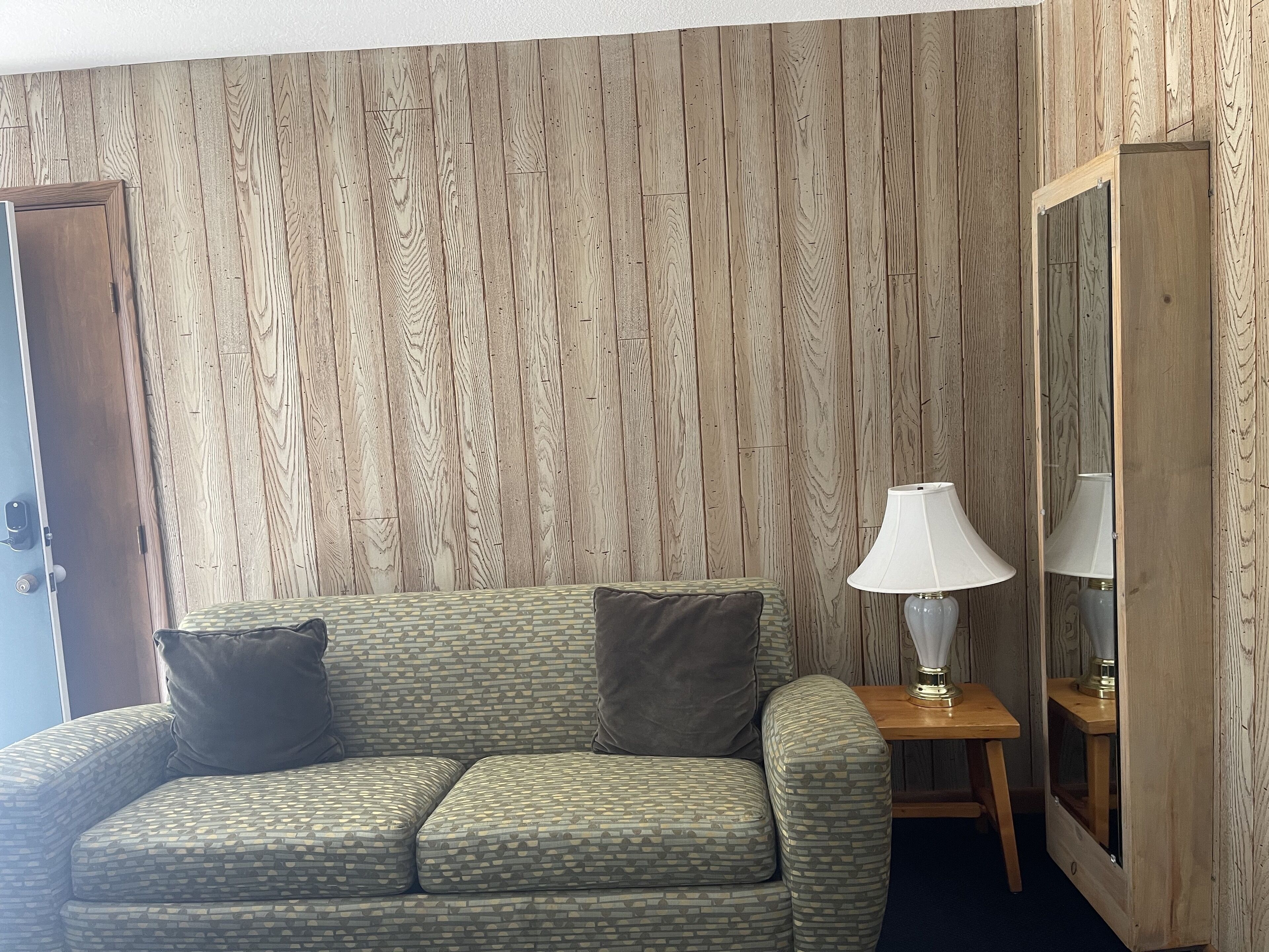 Ein Zimmer mit einer holzverkleideten Wand, einem grünen Sofa mit Kissen und einem kleinen Holztisch mit einer Lampe, sowie einem Spiegel an der Wand.