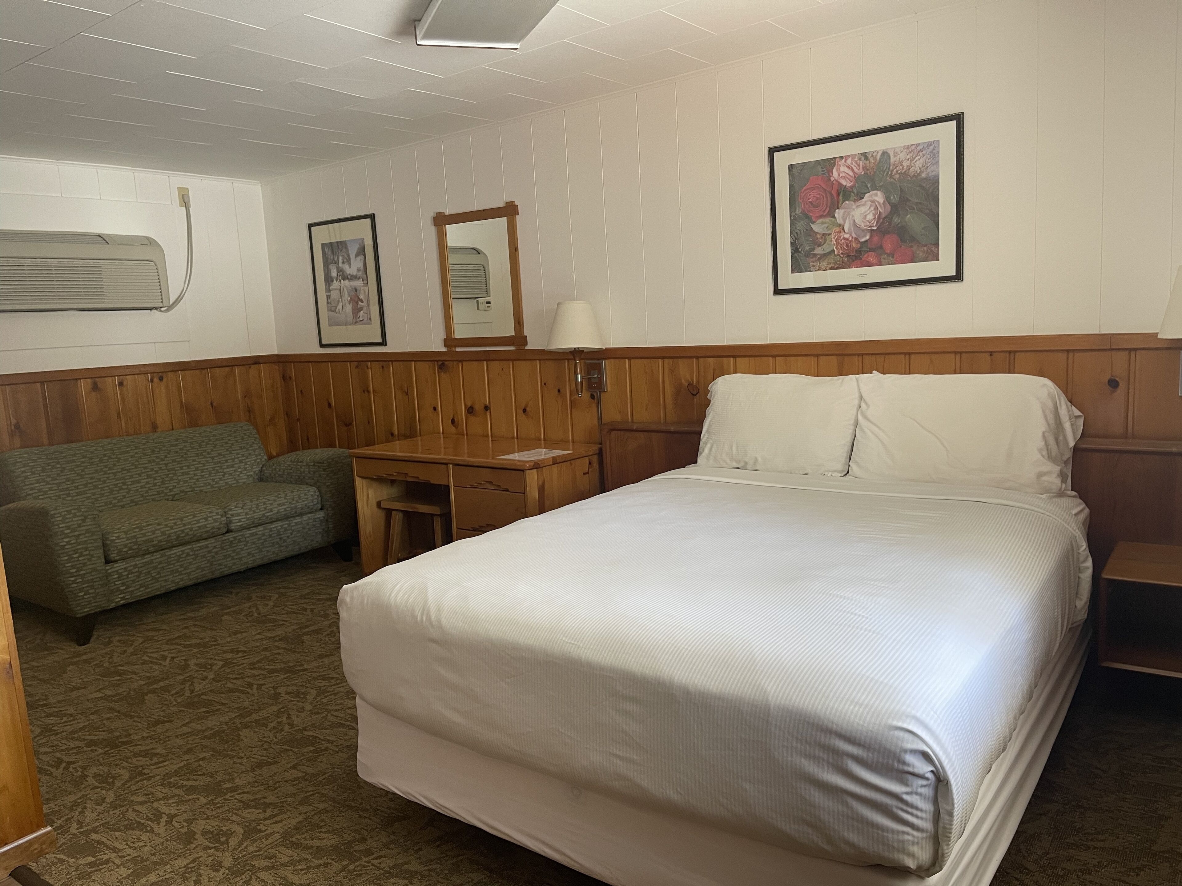 A hotel room with a bed, a desk, a chair, and a wall-mounted air conditioner.