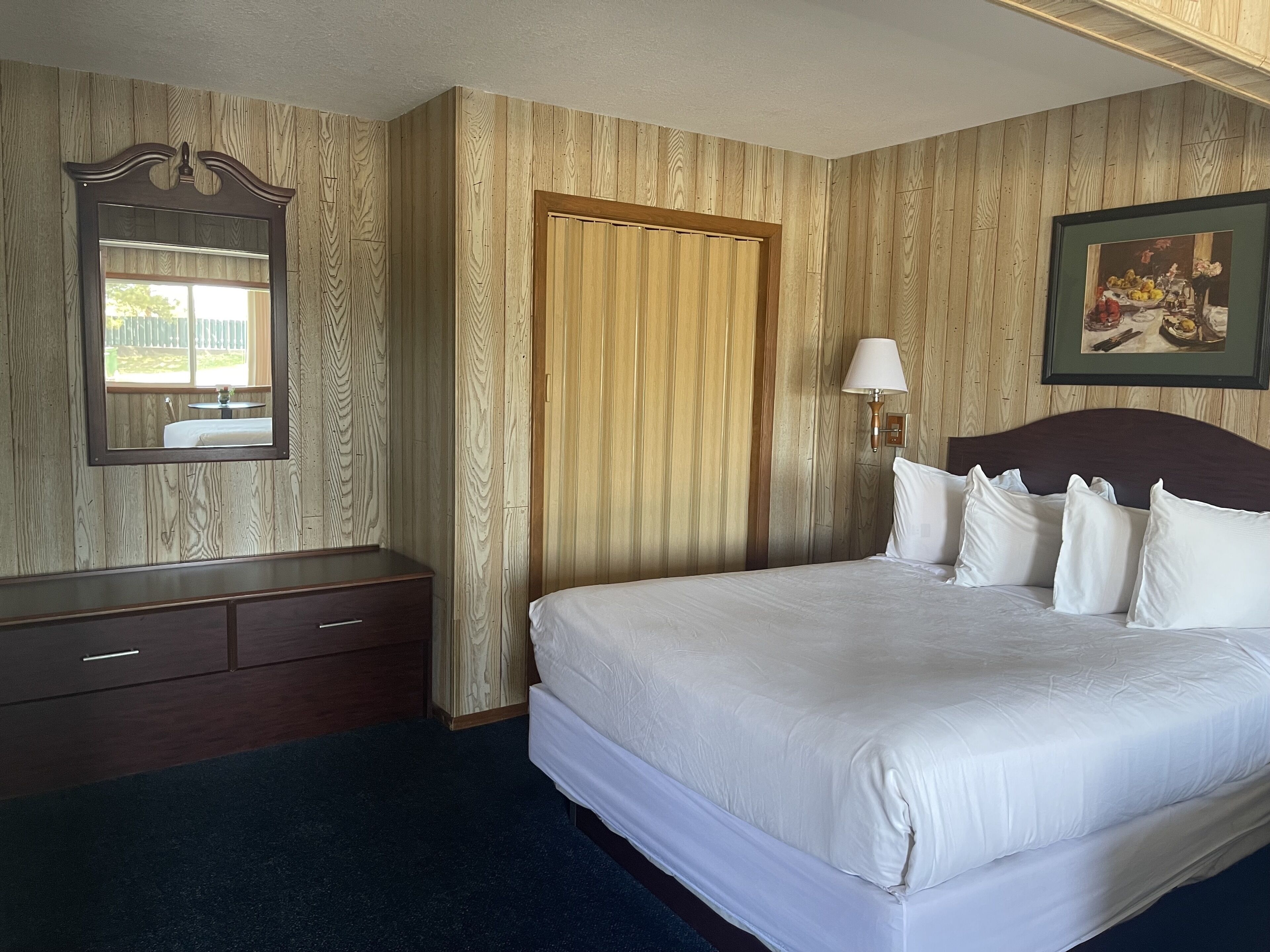 A hotel room with a large bed, a wooden wardrobe, a mirror, a door, and a framed picture on the wall.
