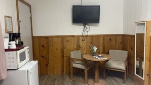 Een kleine kamer met een houten wandpaneel, een aan de muur bevestigde televisie, een magnetron, een koelkast en twee stoelen rond een tafeltje.