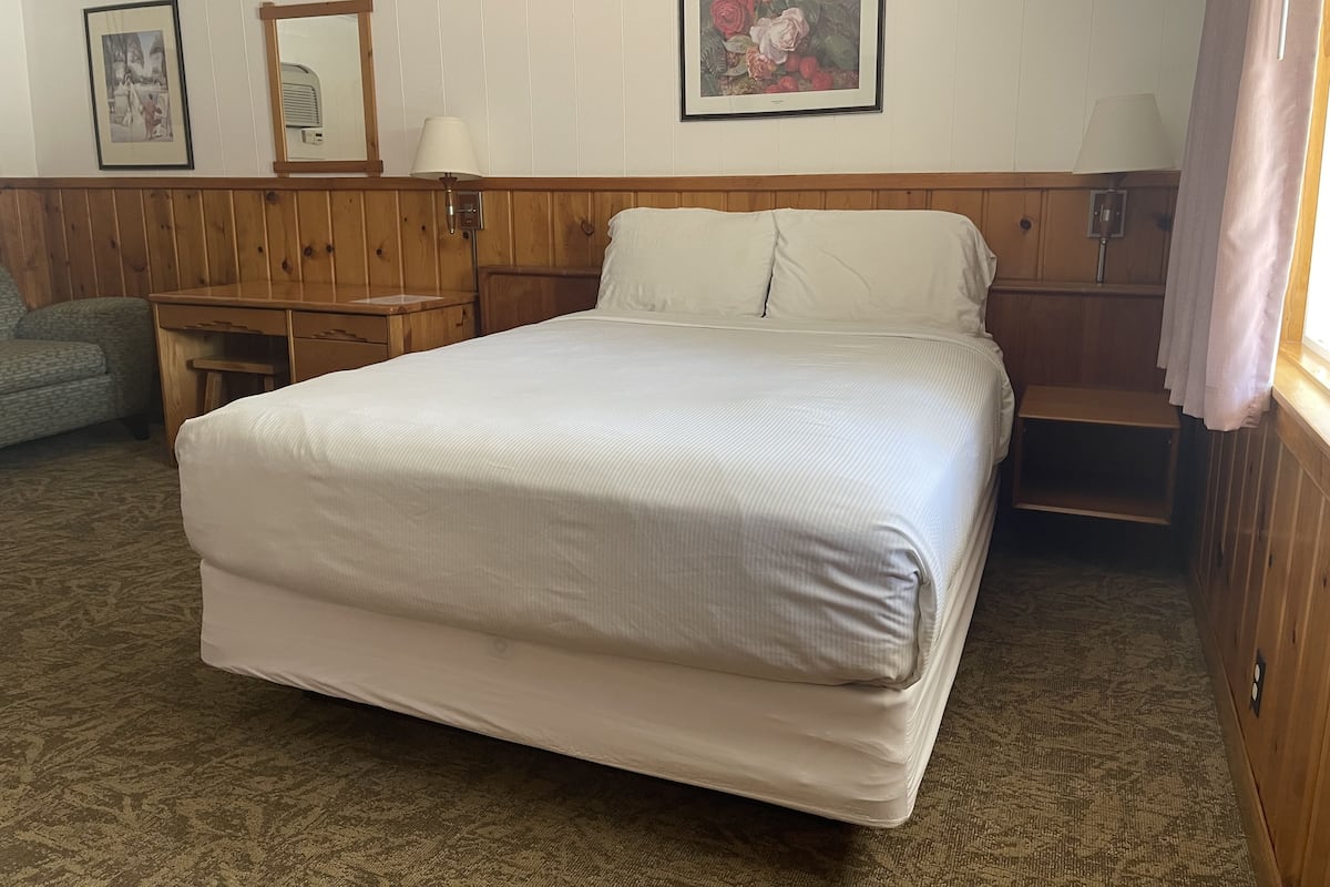 A hotel room with a large bed, wooden headboard, a desk, and a chair. There are framed pictures on the wall and a window with curtains.