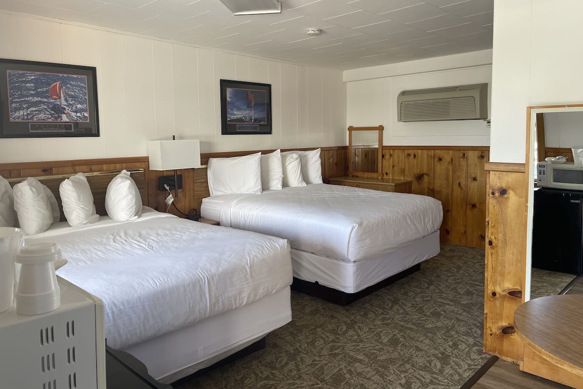 A hotel room with two single beds, a wooden headboard, a bedside table with a lamp, a small mirror, and a microwave in the corner.