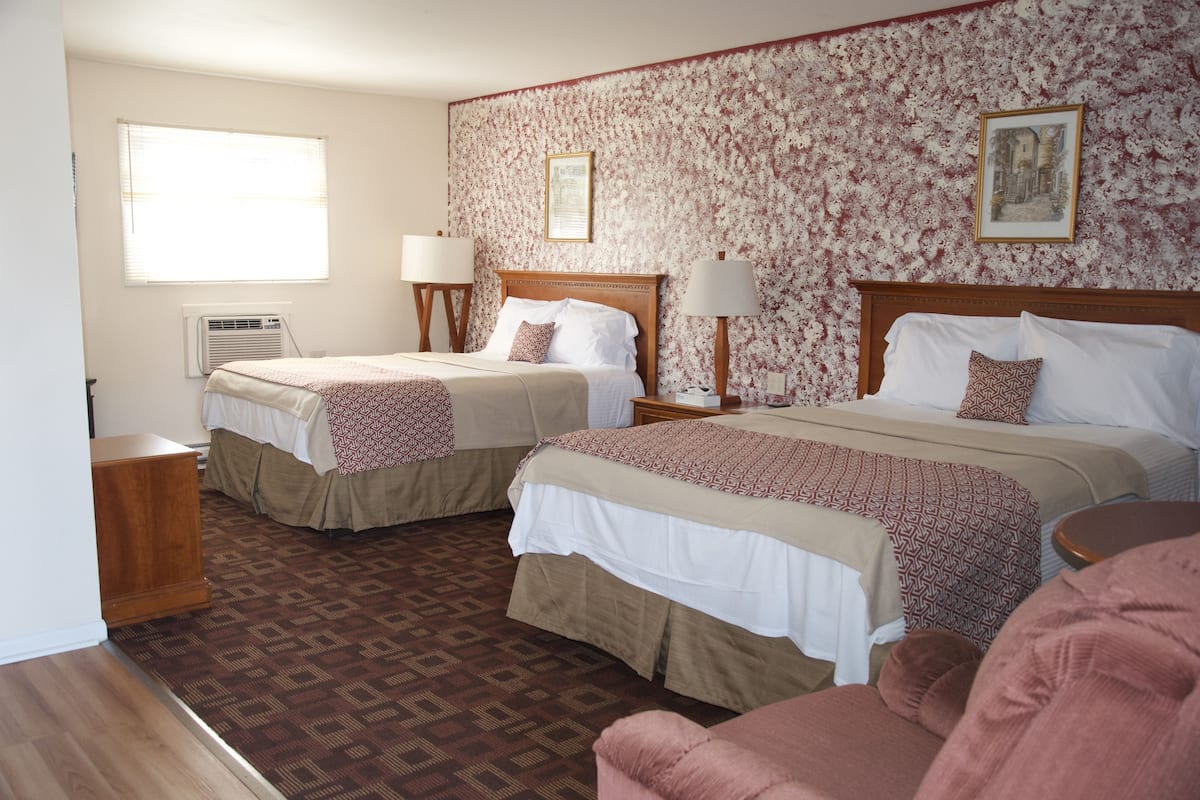 A hotel room with two beds, a wooden headboard, a patterned carpet, and a window with blinds.