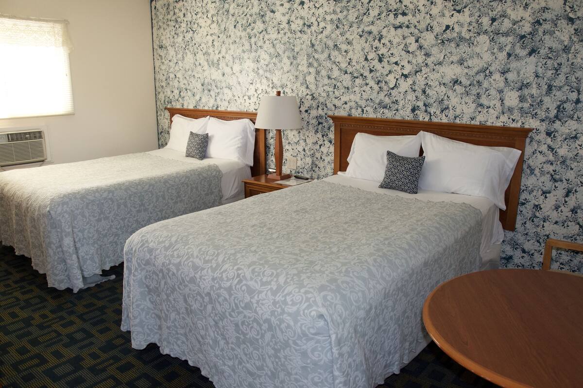 A hotel room with two single beds, a round wooden table, a nightstand with a lamp, and a floral-patterned wall.