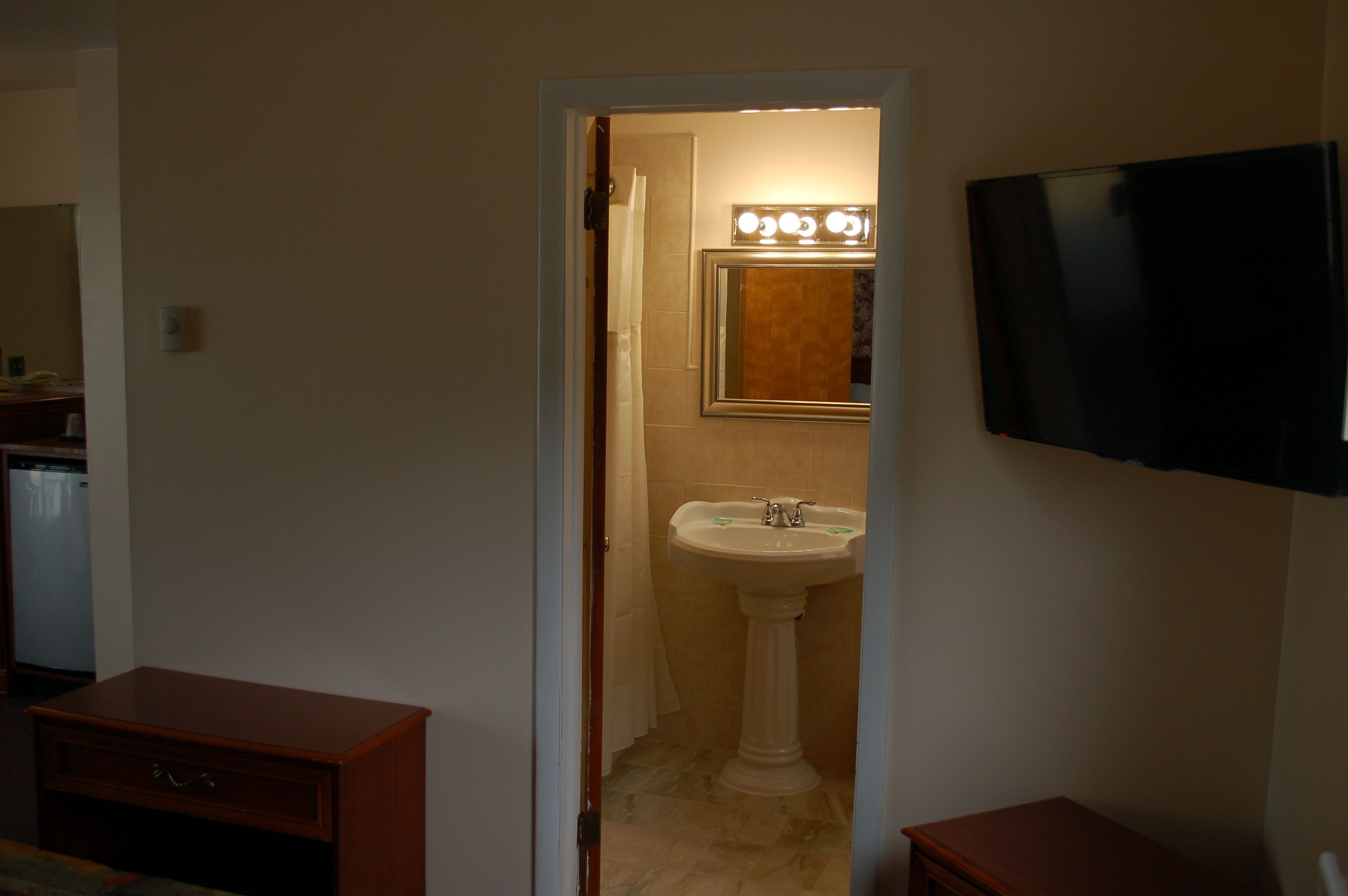 A bathroom with a pedestal sink, a mirror with lights, a television on the wall, and a wooden cabinet.