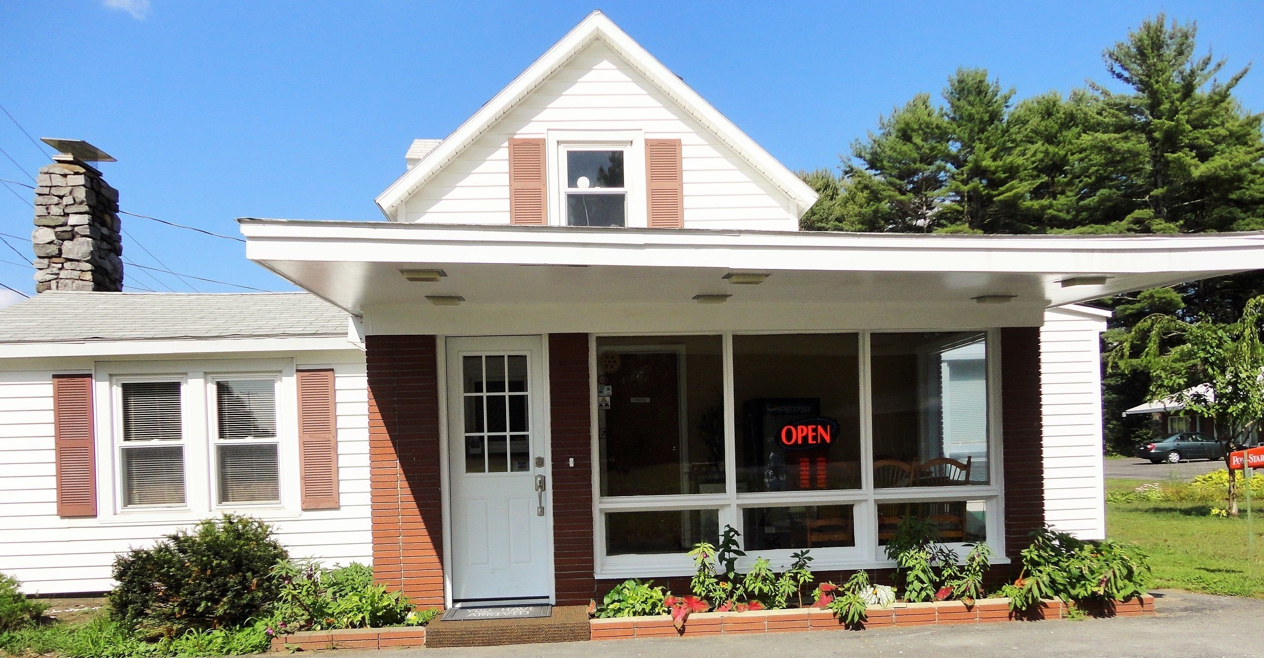 A two-story building with a white facade and brown shutters, featuring a large window with an 'OPEN' sign, a stone chimney, and a small porch area.