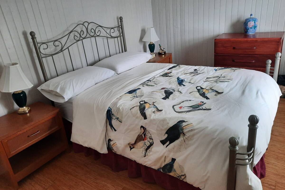 Una cama bien hecha con una colcha blanca con motivos de pájaros, una mesita de noche de madera con una lámpara y un aparador de madera con un jarrón.