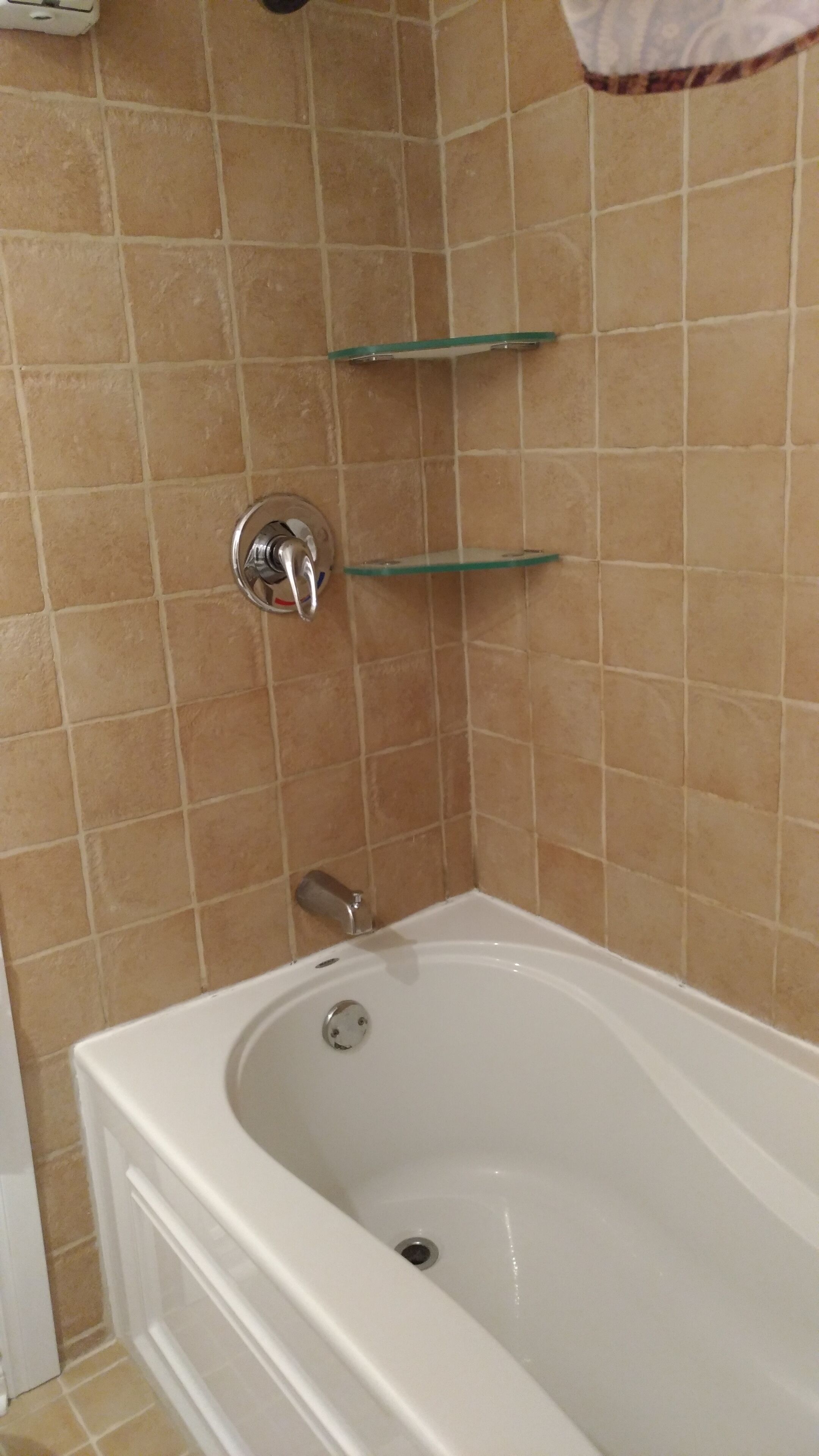 A bathroom with a white bathtub, beige tiled walls, and two glass shelves mounted on the wall.