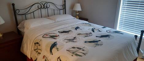 A bed with a white bedspread featuring bird illustrations, a metal headboard, a bedside table with a lamp, and a window with blinds.