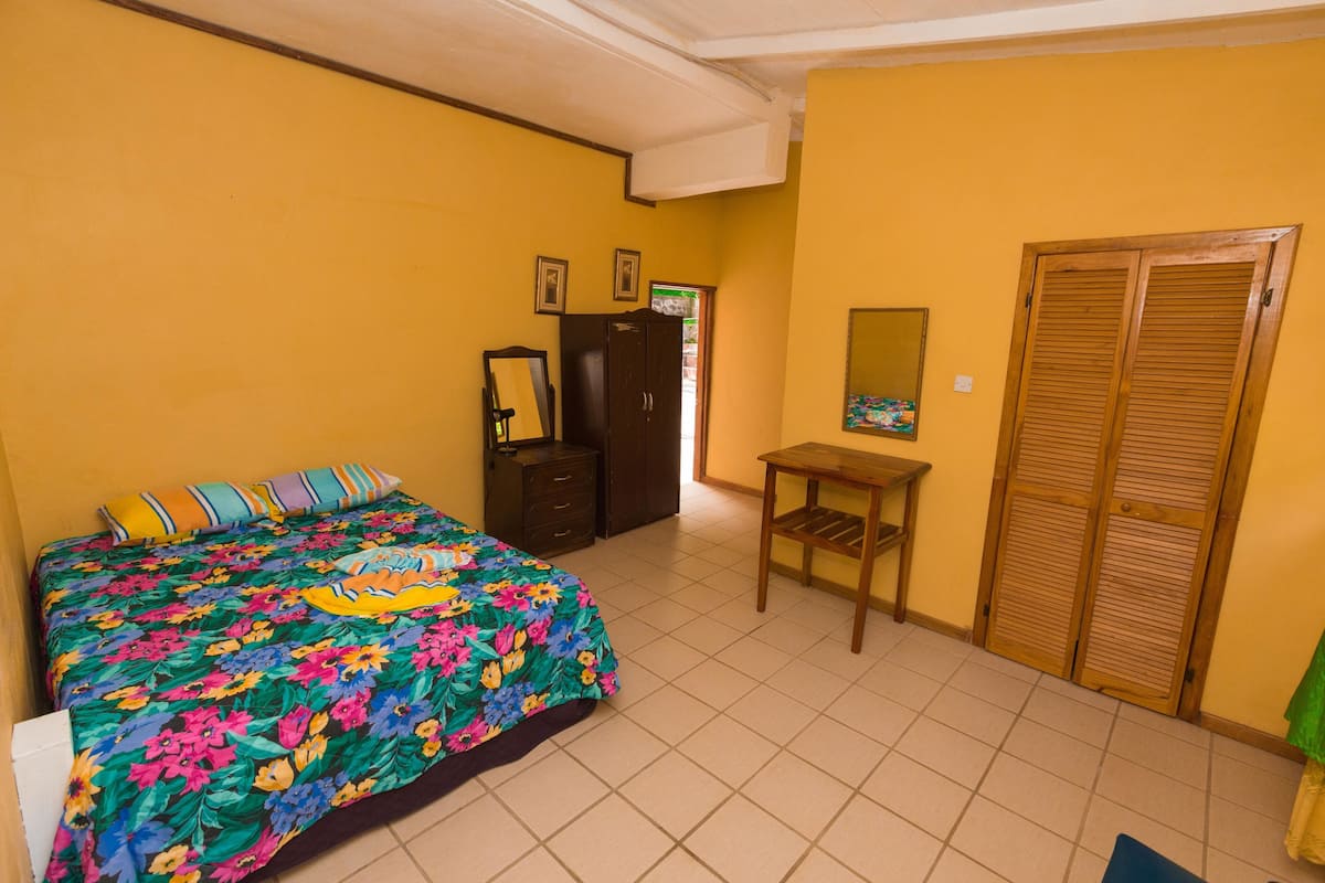 A bedroom with a floral bedspread, a wooden wardrobe, a small table, and a mirror on the wall.