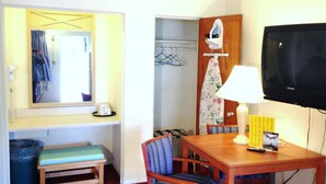 Une chambre d’hôtel avec une table en bois, deux chaises, un banc, une télévision, un miroir, une fenêtre avec des rideaux, un porte-manteau et une porte de salle de bain.