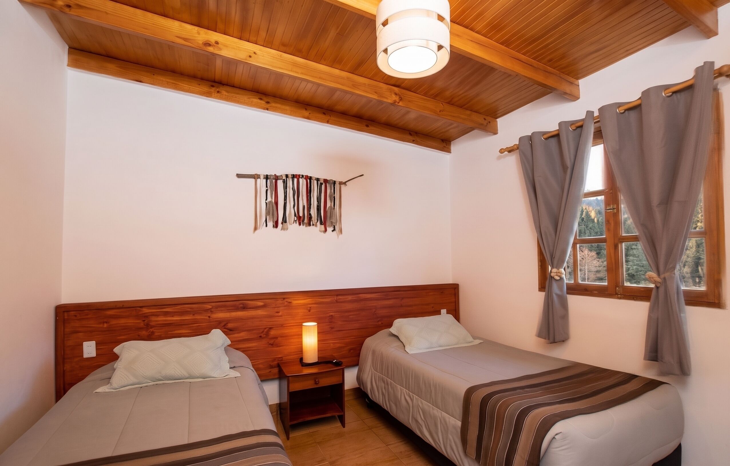 Une chambre avec deux lits, une tête de lit en bois, une table de chevet avec une lampe, une étagère fixée au mur et une fenêtre avec des rideaux.