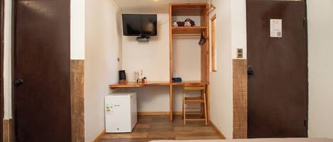 Habitación de hotel compacta con cama, escritorio con computadora, refrigerador pequeño y televisión montada en la pared.