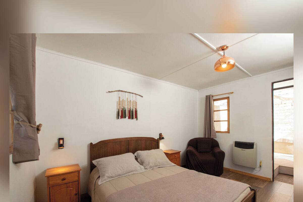 Un dormitorio con cama, mesitas de noche, ventilador de techo, lámpara de pared, ventana y radiador.