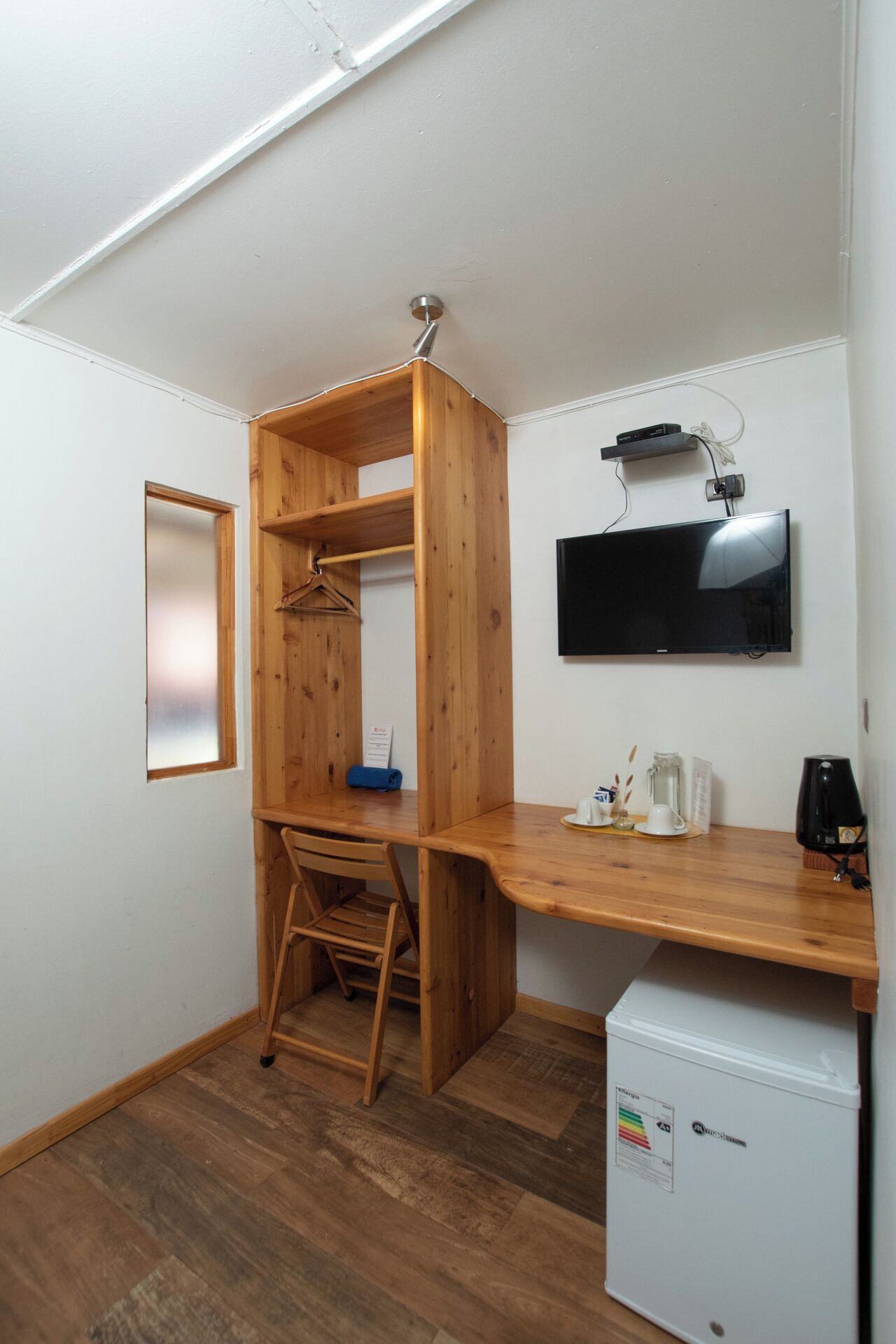 A compact room with a wooden desk, a small refrigerator, a TV mounted on the wall, and a wooden wardrobe with a ladder.