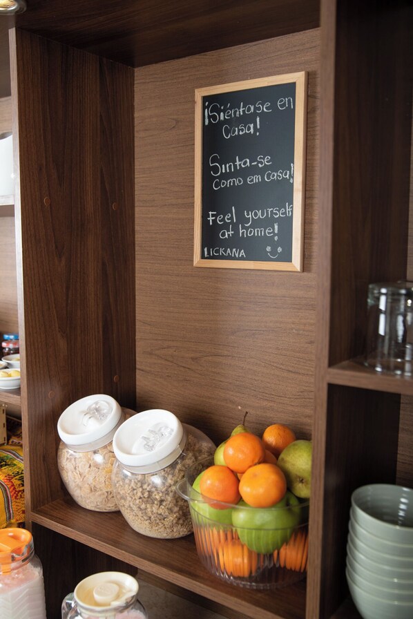 Una estantería de madera con botes de avena y un cuenco de fruta. Un cartel de pizarra dice: "Siéntete como en casa!"