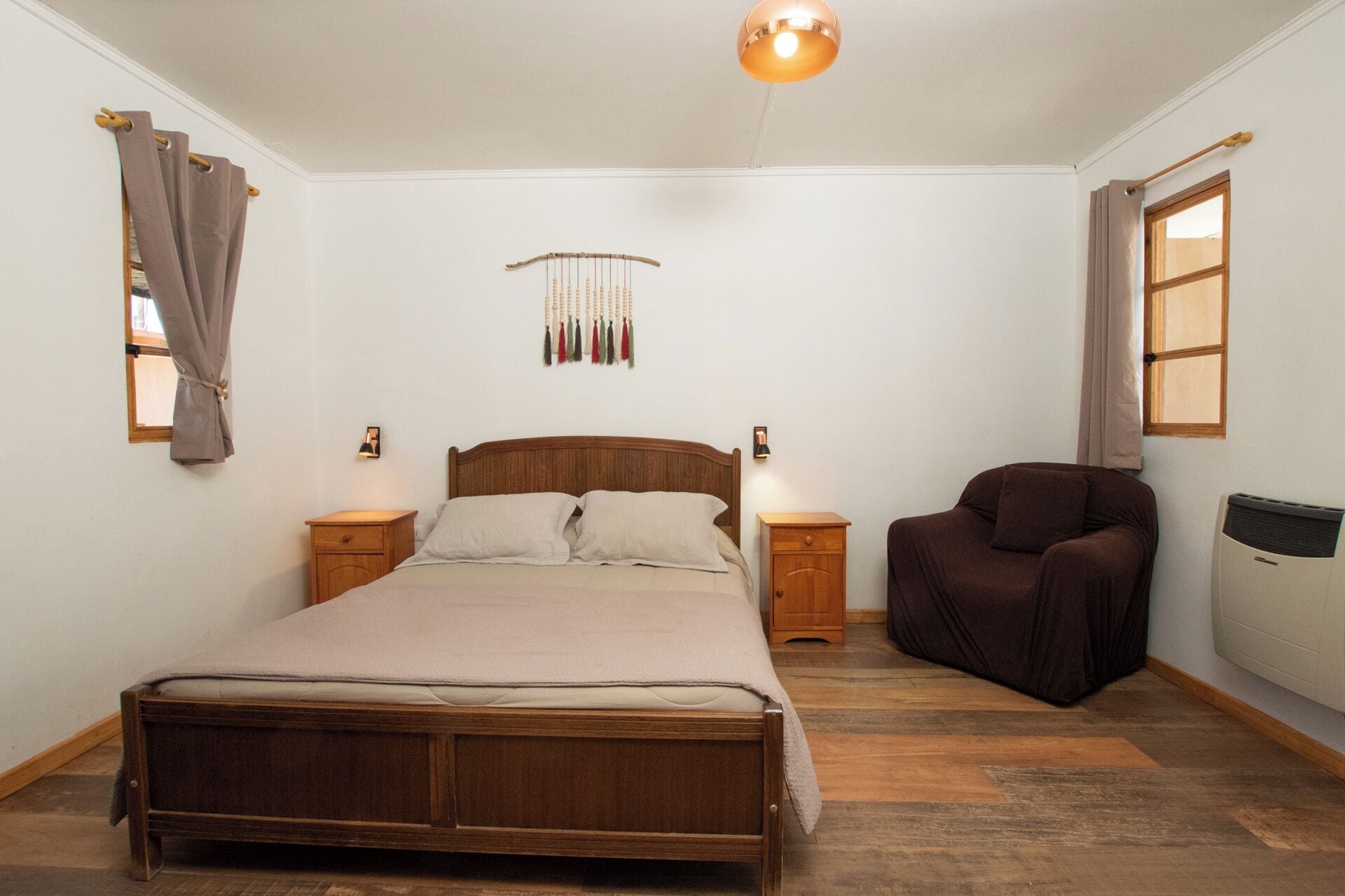 A bedroom with a wooden bed, bedside tables, a chair, a wall-mounted lamp, and a window with curtains.