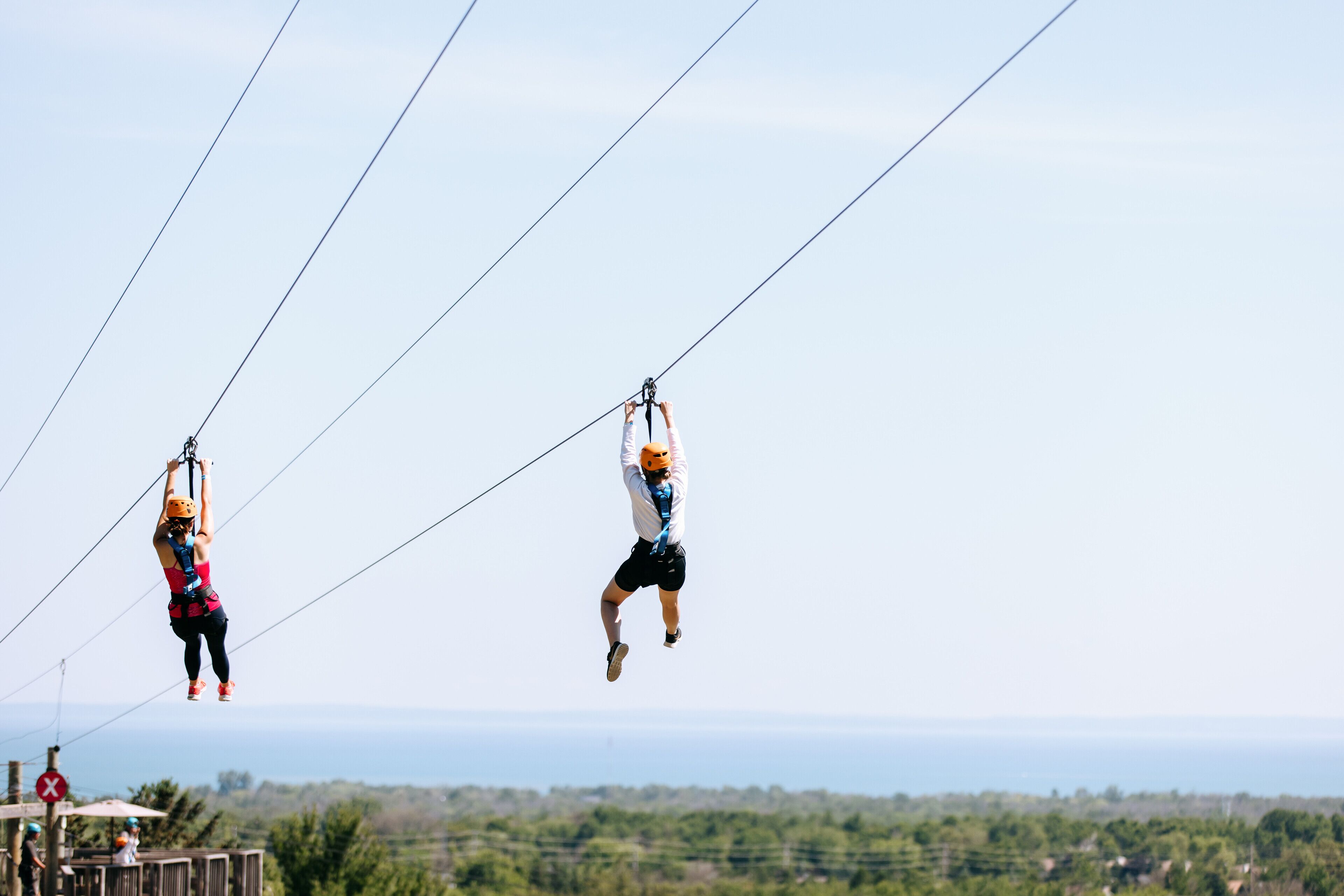 Two people ziplining over a landscape with a view of the ocean.