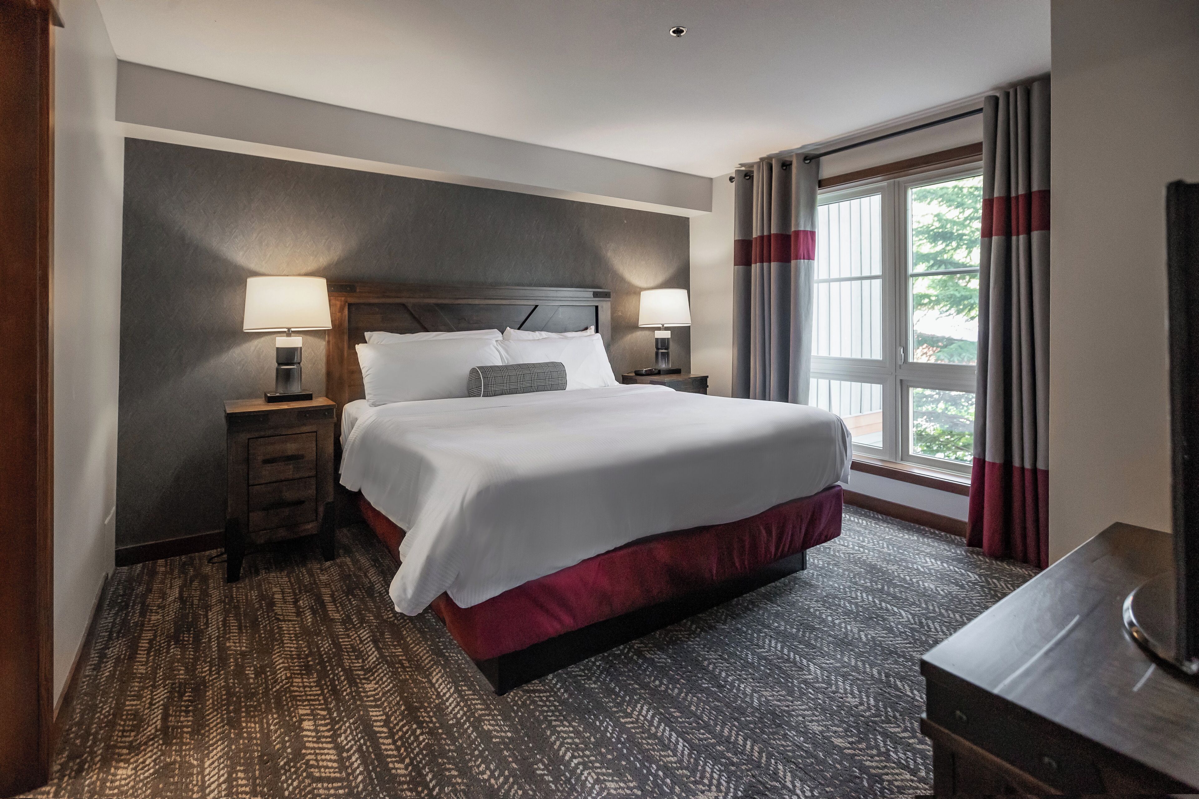 A hotel room with a large bed, two bedside tables with lamps, a wooden headboard, a large window with curtains, and a patterned carpet.