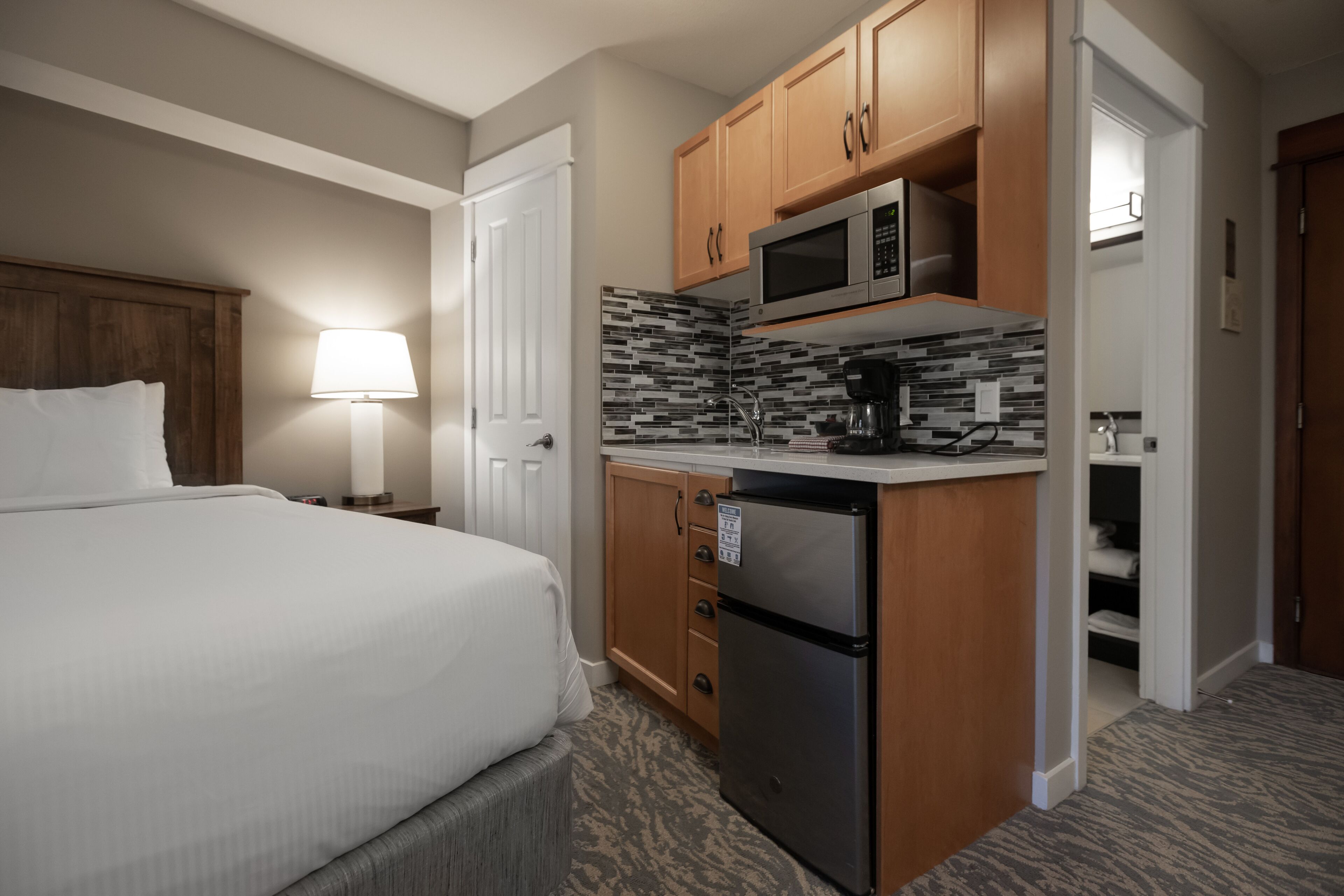 A hotel room with a bed, a desk with a microwave, a refrigerator, and a tiled backsplash.